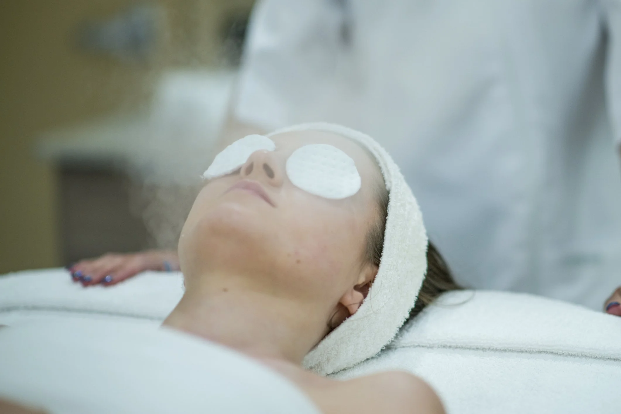 A woman is lying on a spa bed with white towels covering her forehead and eyes, receiving a steam treatment in a spa or beauty clinic.