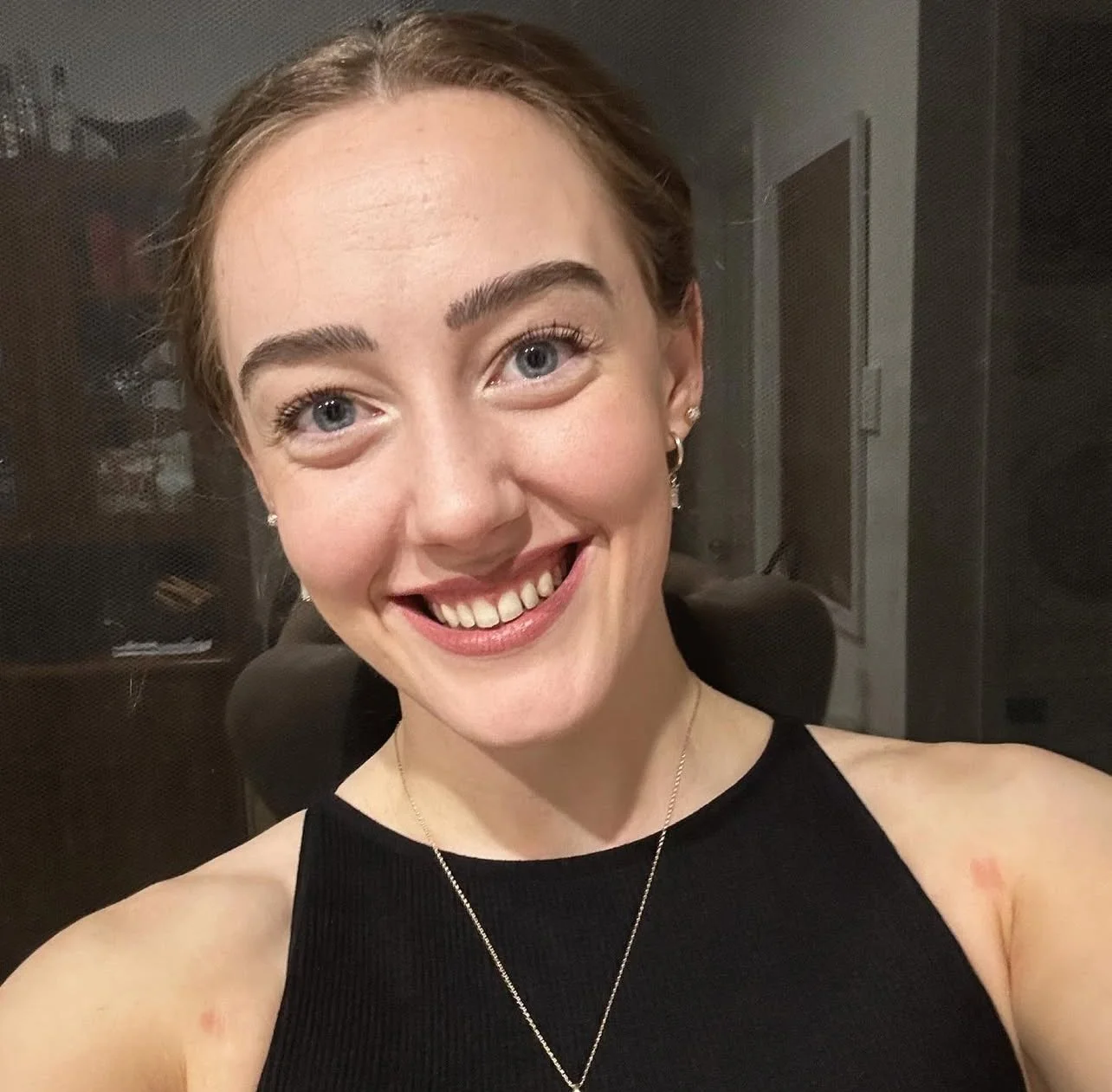 A close-up photo of a smiling woman with blue eyes, light skin, and brown hair, wearing a black sleeveless top, earrings, and a delicate necklace. The background appears to be a room with a dark wall and some furniture.