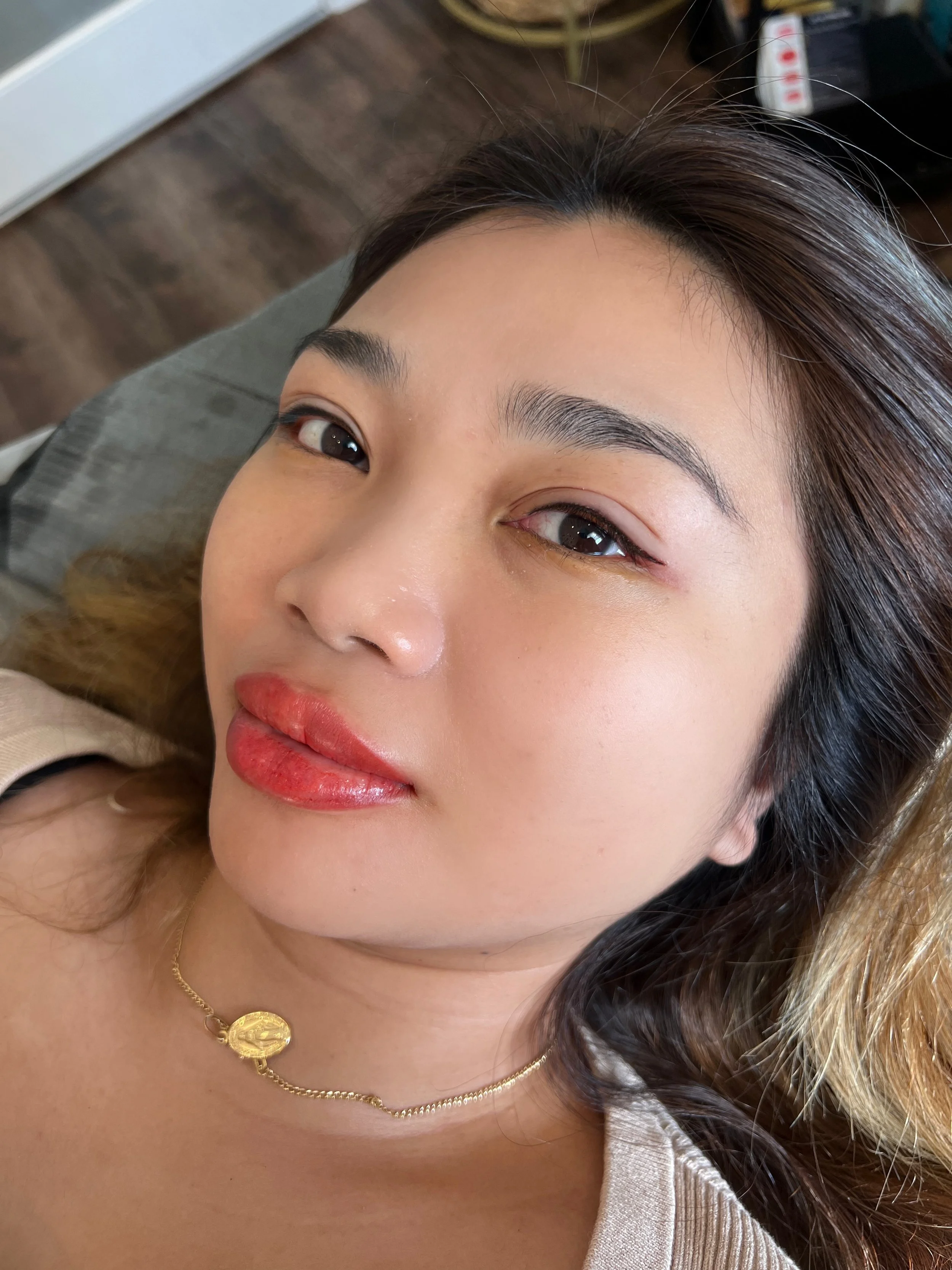 A woman with long dark hair and makeup, lying down and looking at the camera, wearing a gold necklace