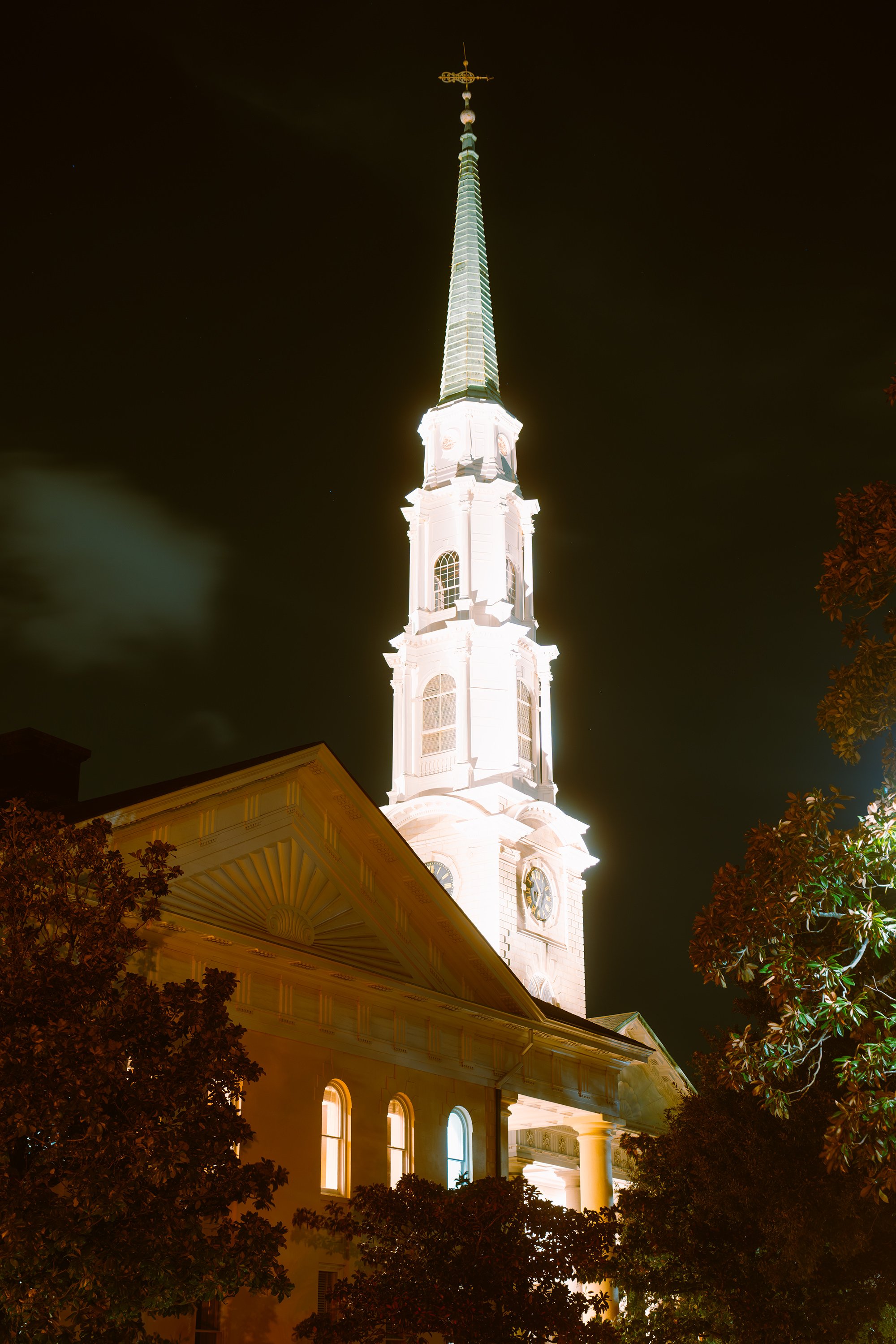 Night view of a tall, illuminated church steeple with a golden cross at the top, surrounded by trees.