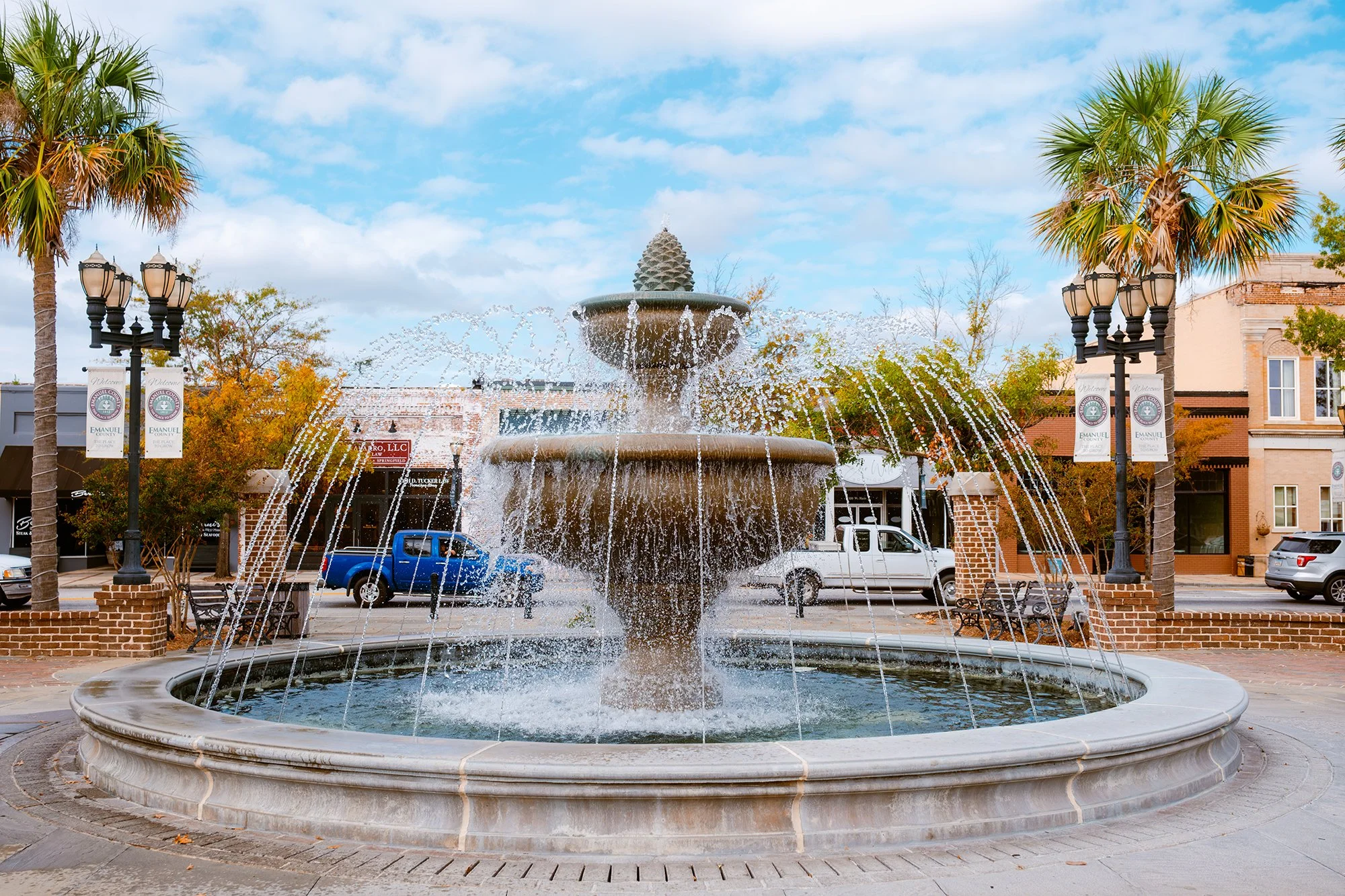 Public square with ornate fountain, palm trees, and benches under a partly cloudy sky.