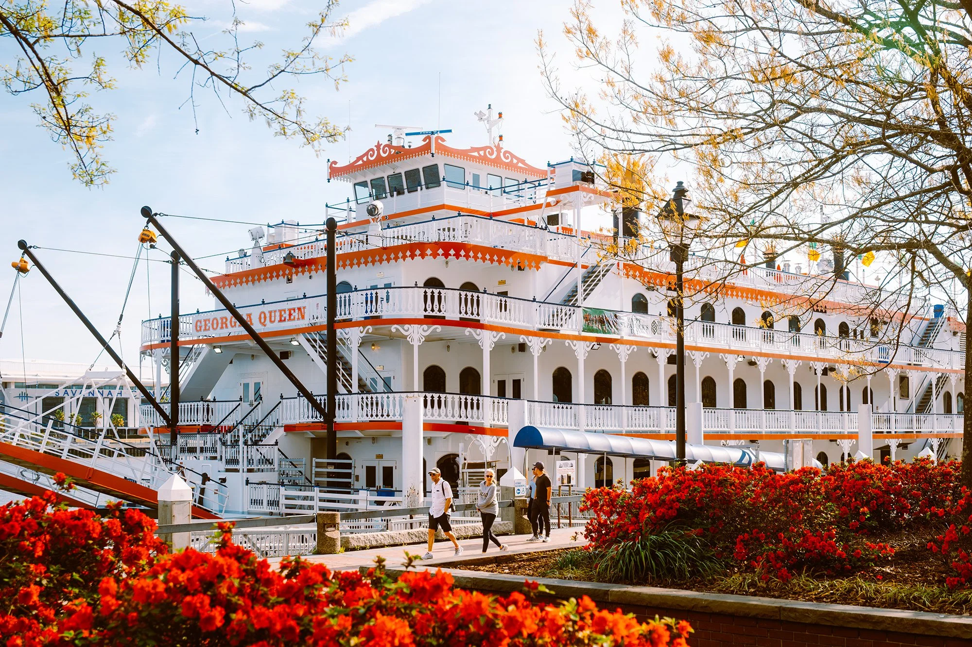 The Savannah Riverboat, called Georgia Queen, docked at a pier with people walking nearby, surrounded by blooming red flowers and trees with some leaves falling.