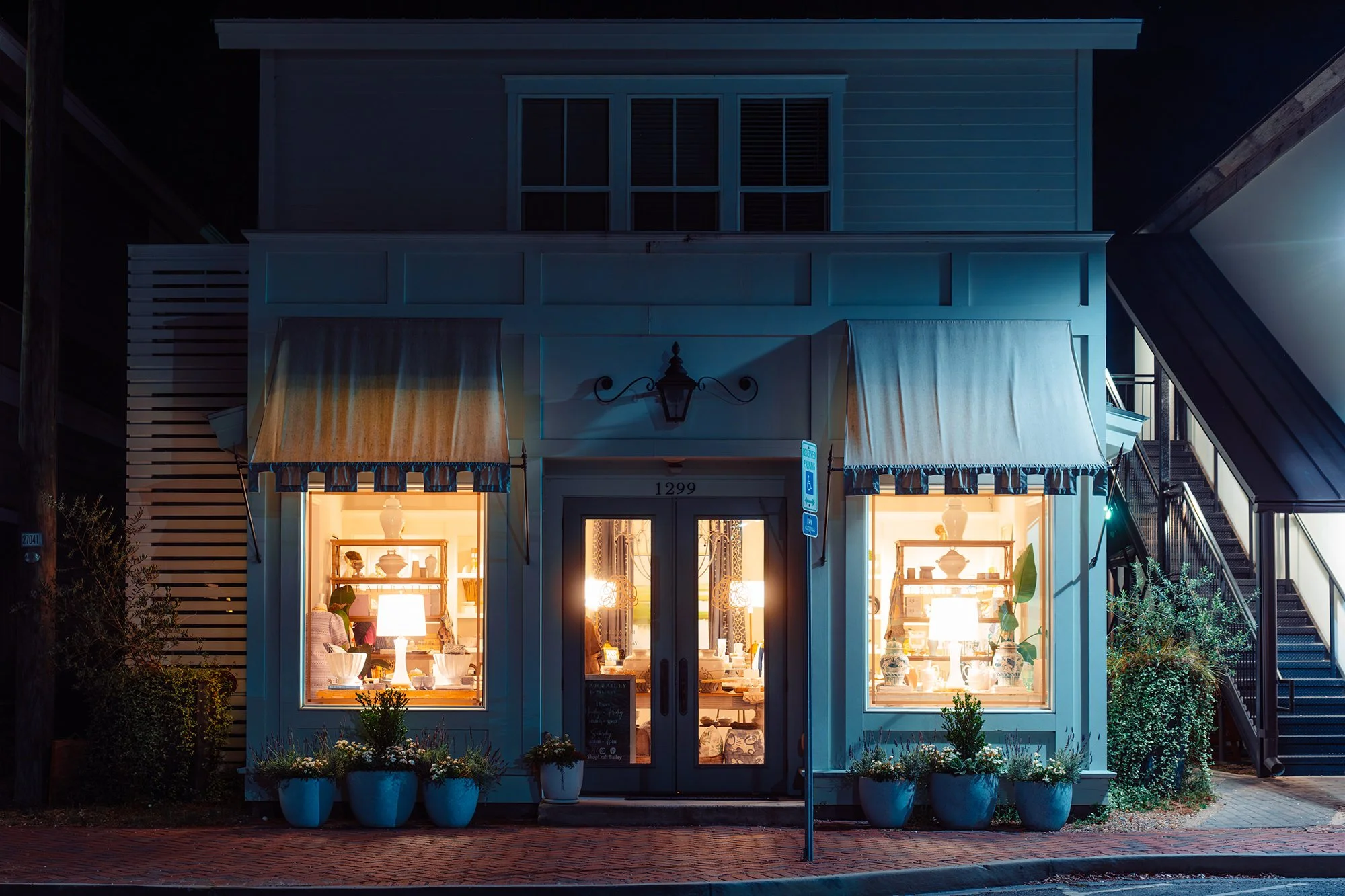 A storefront shop at night with large illuminated display windows showcasing various decorative items and lamps. Potted plants line the sidewalk in front and an sign stands near the entrance.
