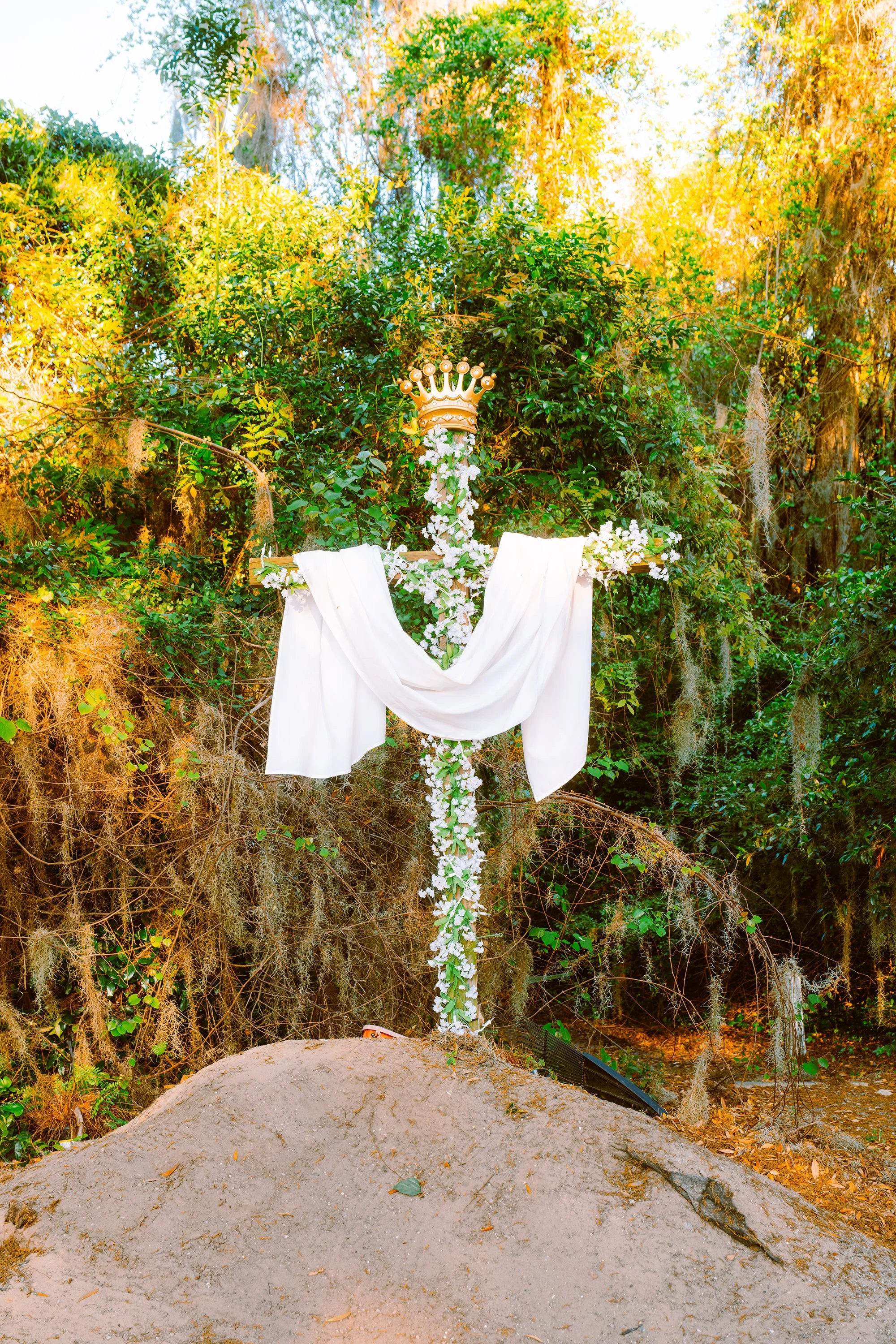 A decorated cross with a gold crown top, white drapery, and floral arrangements stands on a mound of dirt in a wooded area, with sunlight filtering through trees.