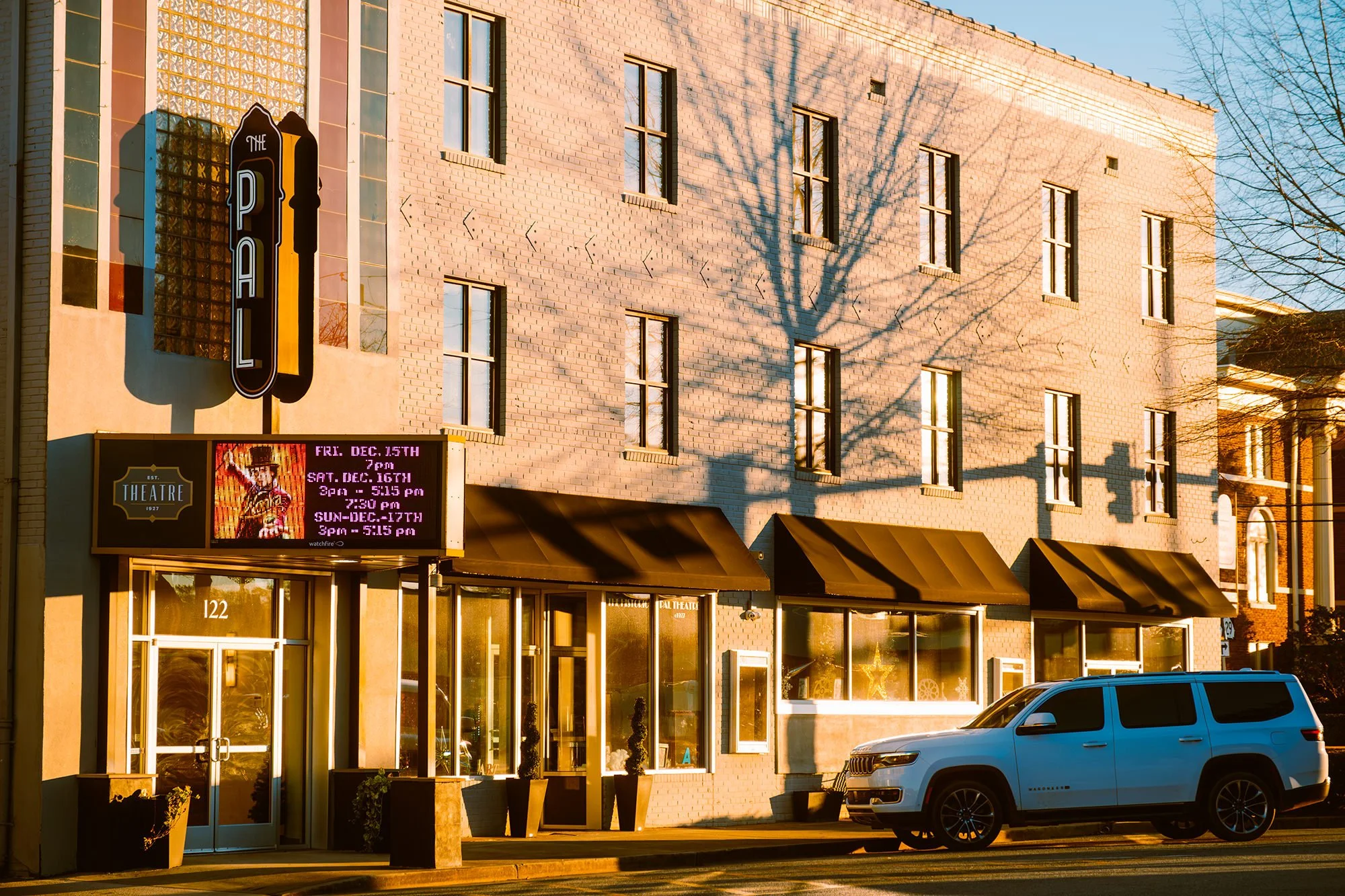 Street view of a theater with a marquee, a white SUV parked in front, and a brick building with multiple windows. The marquee displays upcoming showtimes, and there is a vertical sign with the word "PAL". The building has three smaller square windows