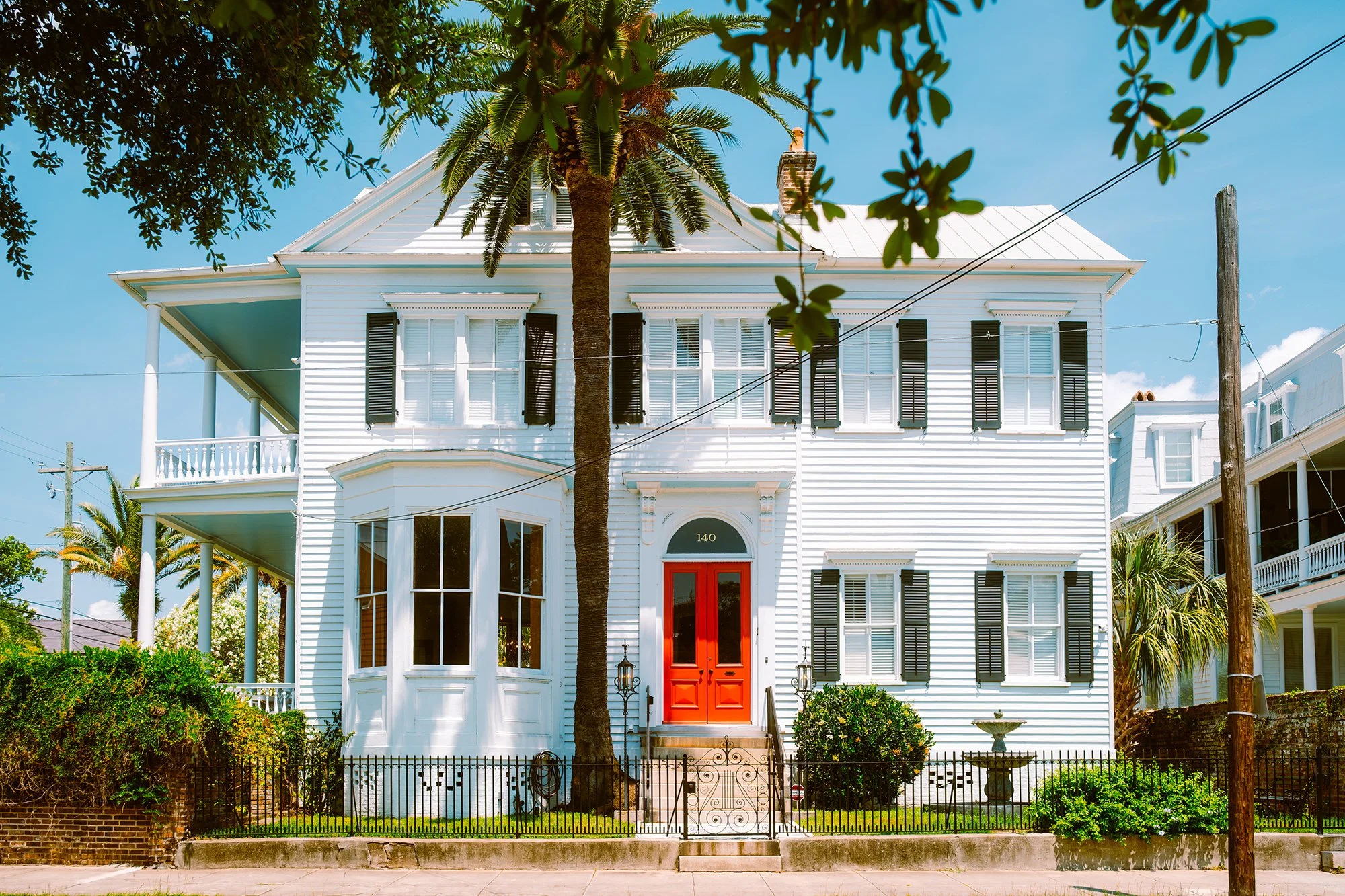 A large white house with black shutters and a red front door, surrounded by a small yard with a tree in front, a fountain, and bushes, under a blue sky.