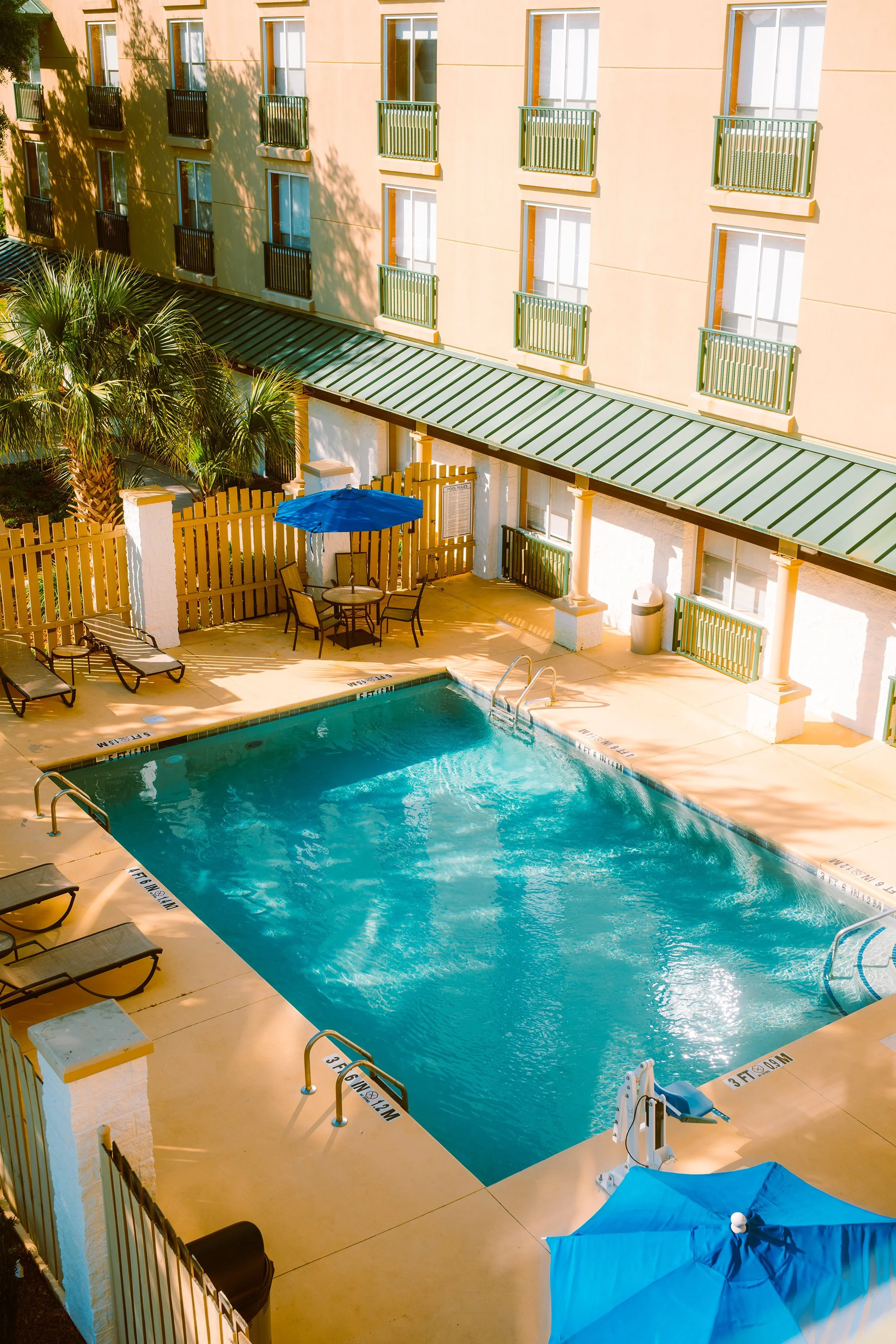 An outdoor hotel pool area with lounge chairs, a table with an umbrella, and a building with multiple windows and balconies in the background.