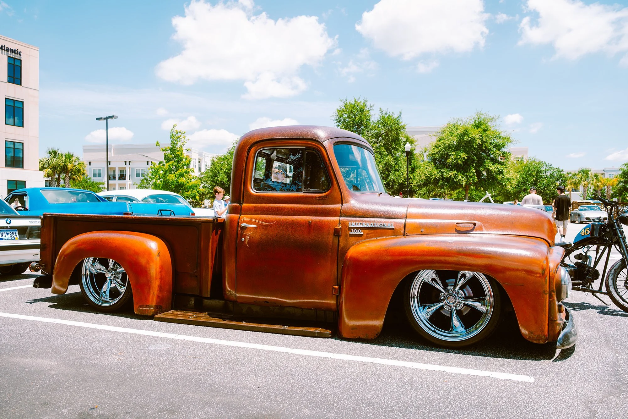 A vintage rusty brown pickup truck with shiny custom wheels parked in a lot during a car show.