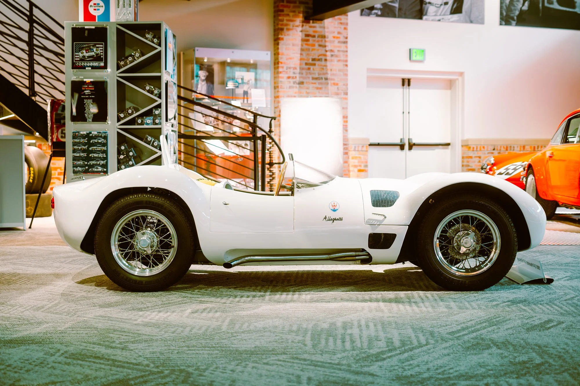 White vintage race car with spoked wheels on display inside a showroom, with other classic cars and automotive memorabilia in the background.