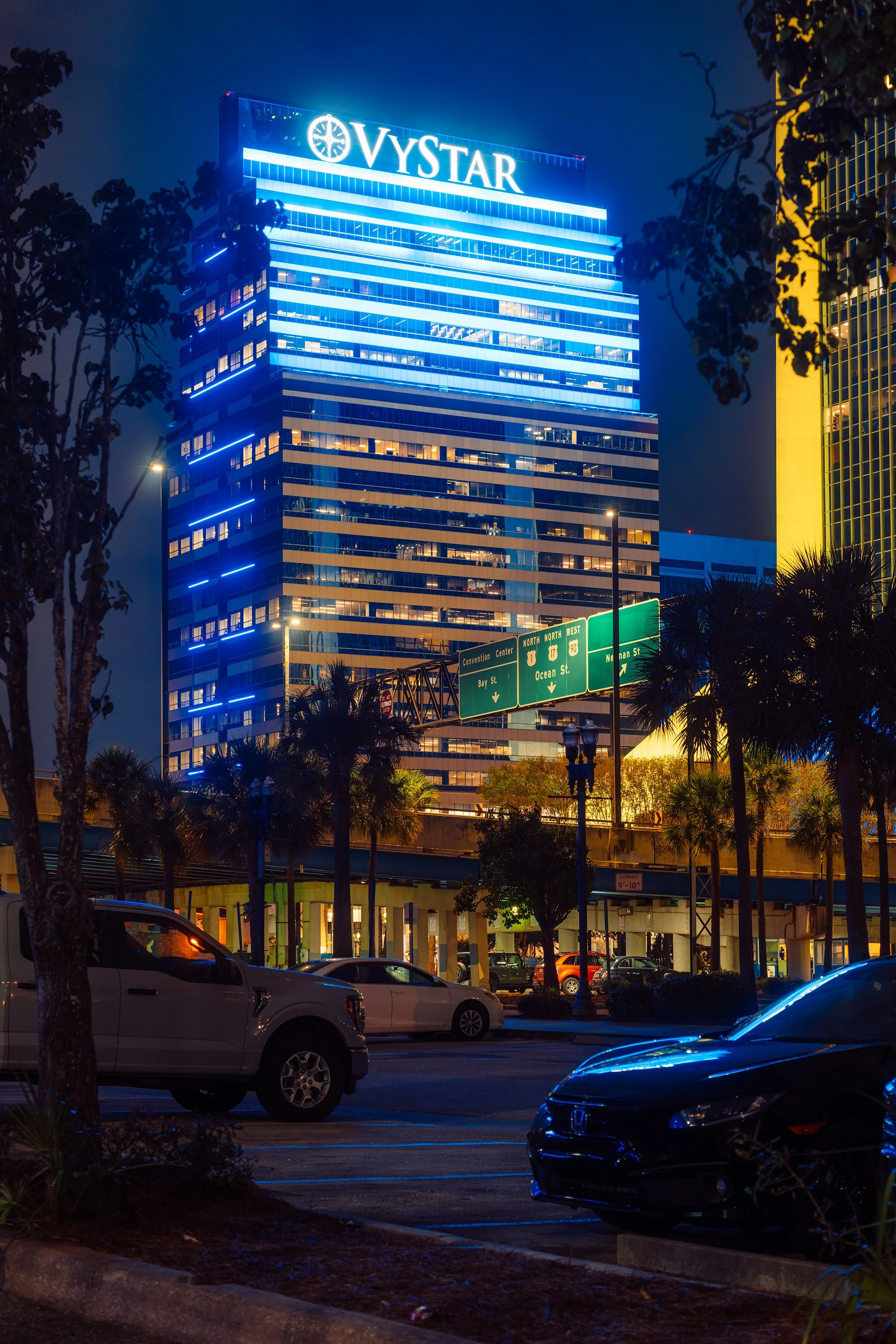 Night view of a tall glass office building illuminated with blue and yellow lights, featuring the sign 'VYSTAR' at the top, with palm trees, cars parked in the foreground, and green road signs below.