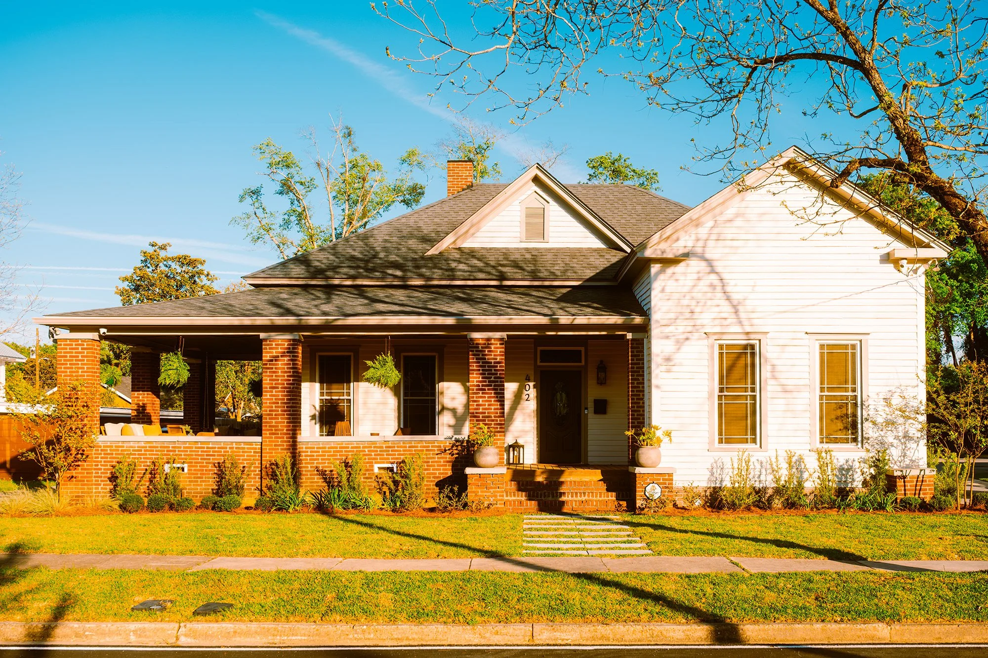 A suburban house with a front porch, brick pillars, a dark roof, white and brick exterior walls, large windows, and a well-maintained lawn with plants and trees.