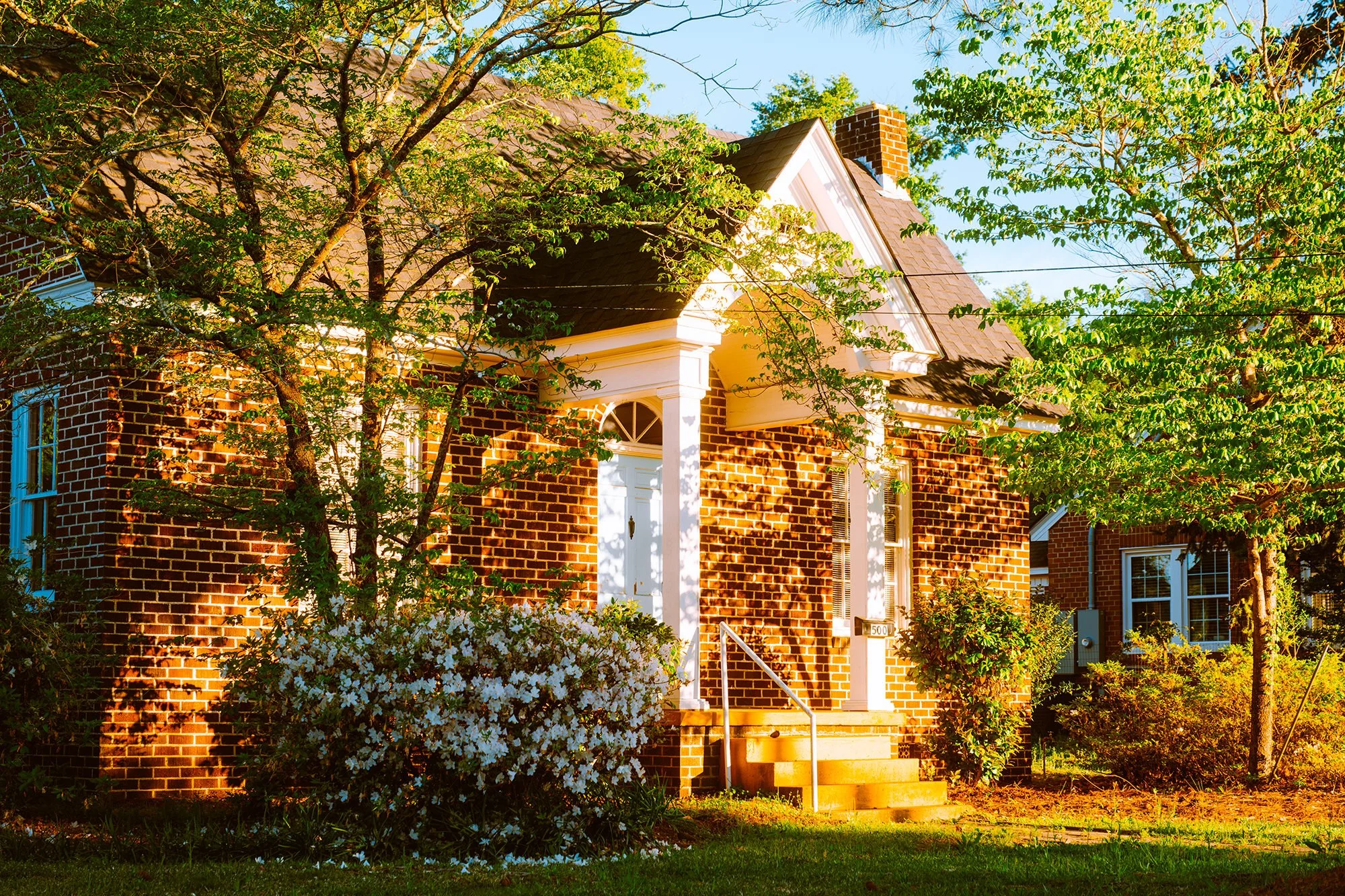 A brick house with a white front porch, steps, and railing. Surrounded by green trees and a flowering shrub in the front yard. Bright sunshine casts shadows on the house in the late afternoon.