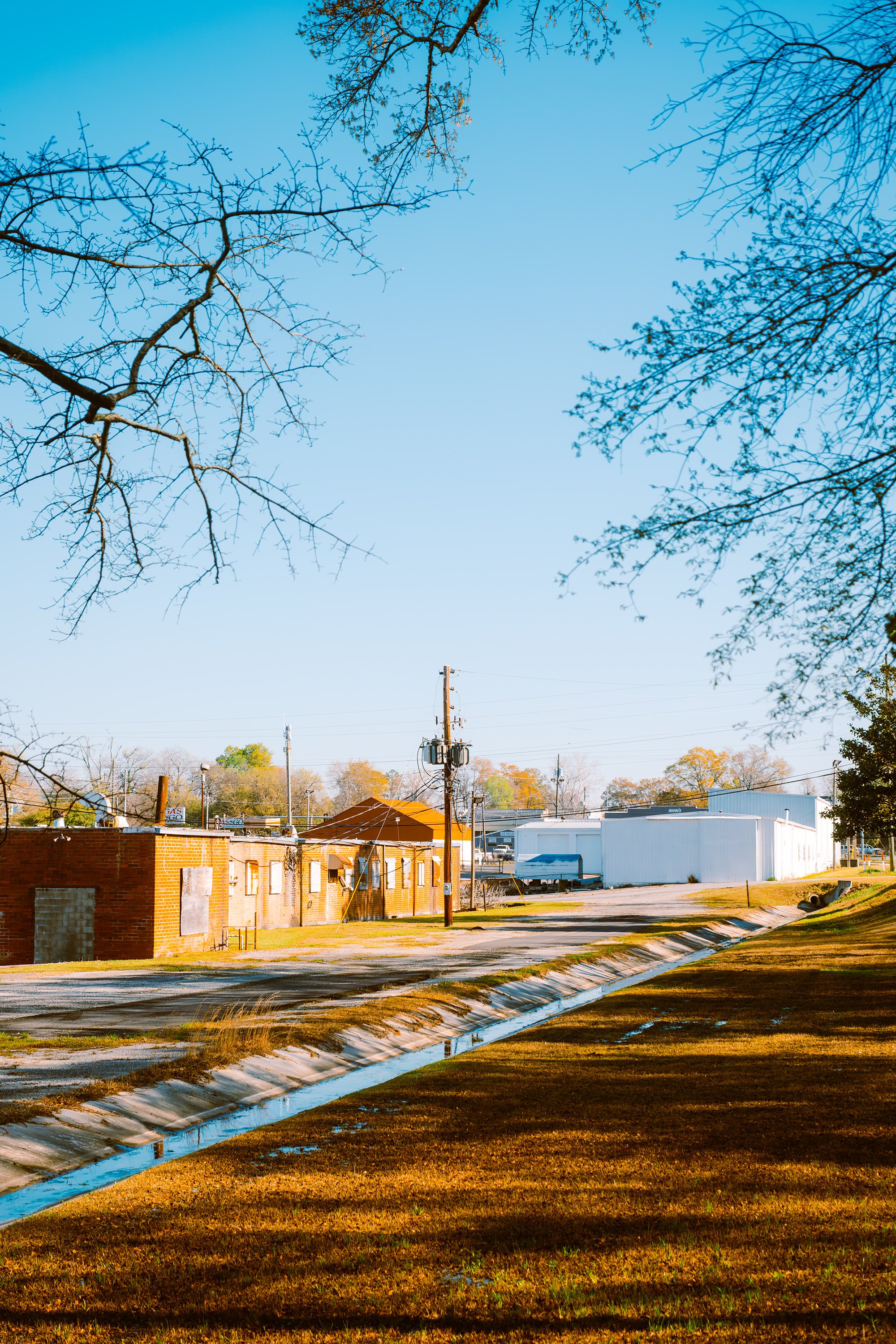 A small town scene with a brick building, a white warehouse, power lines, leafless trees, and a grassy area with a drainage ditch reflecting the sky, under clear blue skies.