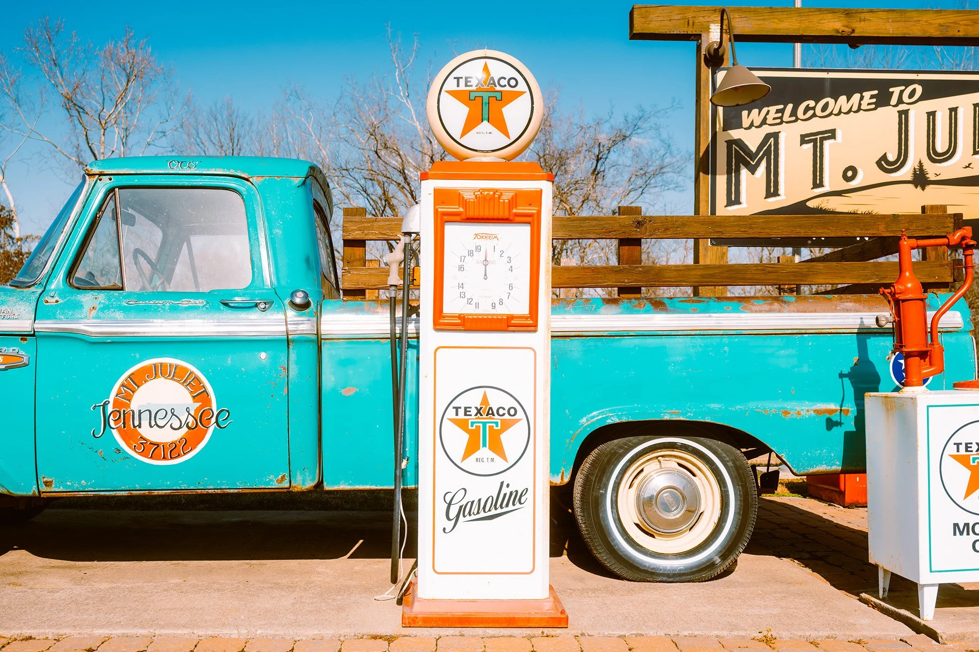 Vintage turquoise pickup truck with rust spots, parked at a Texaco gas station, with a white and orange Texaco gas pump in front of it. A sign to the right reads 'Welcome to Mt. Juliet'.