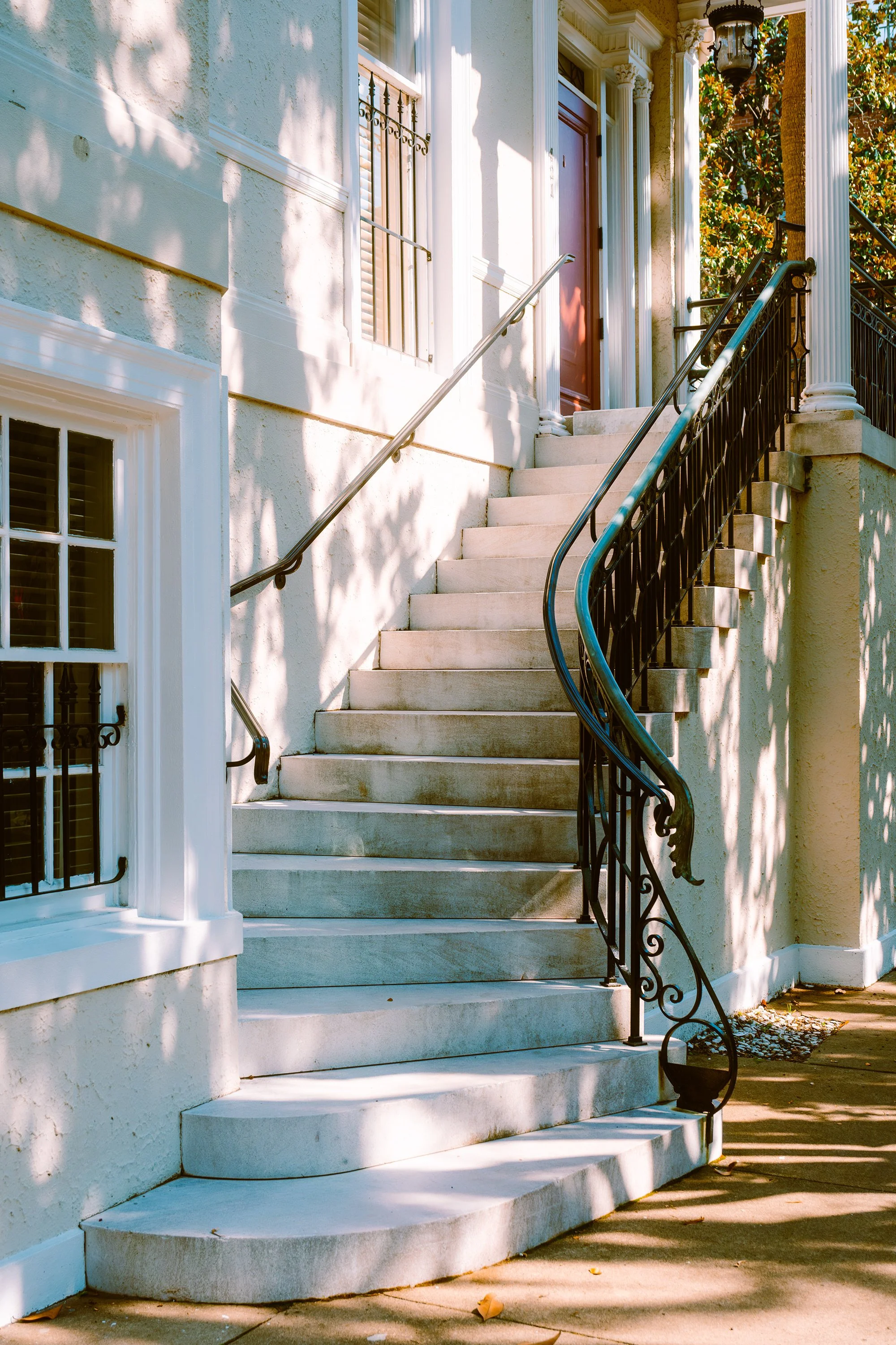 Exterior view of a residential staircase with ornate black railings and a brown front door, in sunlight with shadows and some fallen leaves on the sidewalk.