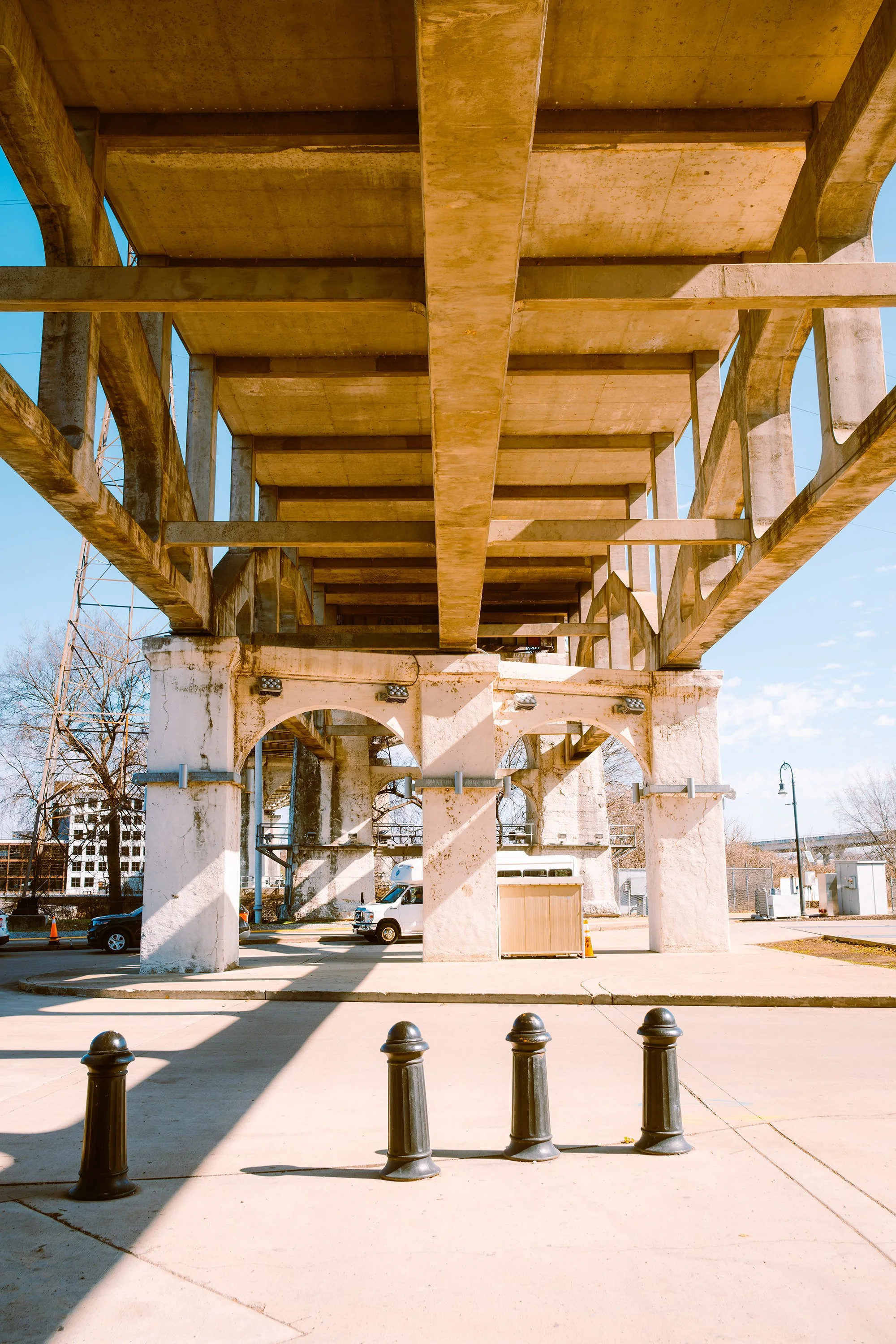 Underneath a bridge structure with concrete columns and wooden beams, a parking lot with a few cars and a utility box, against a clear blue sky.