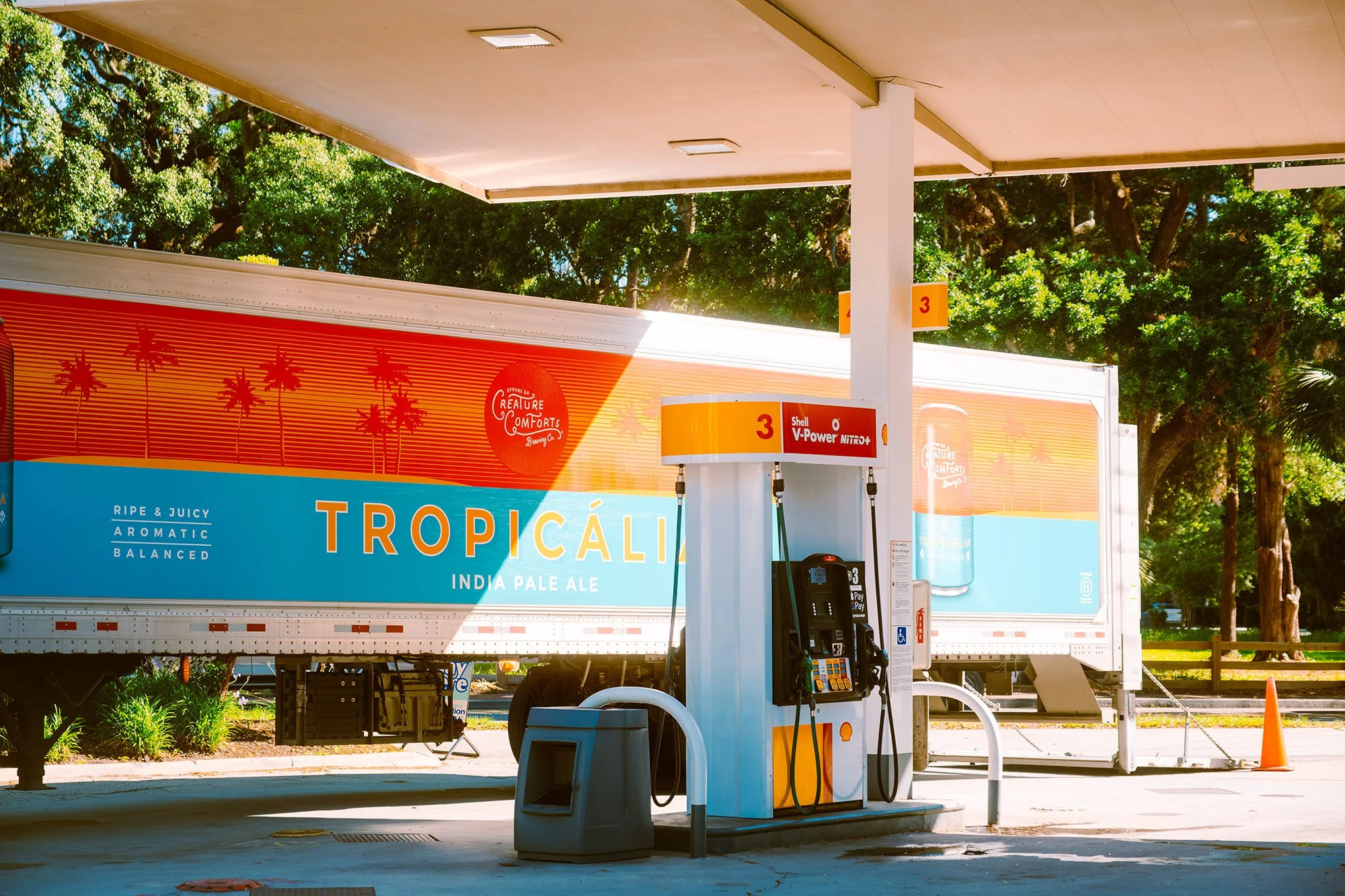 A Shell gas station with a single pump labeled '3' and 'V-Power Nitro+' in front of a large orange and blue trailer advertisement for Tropical India Pale Ale, surrounded by trees and a wooden fence in the background.
