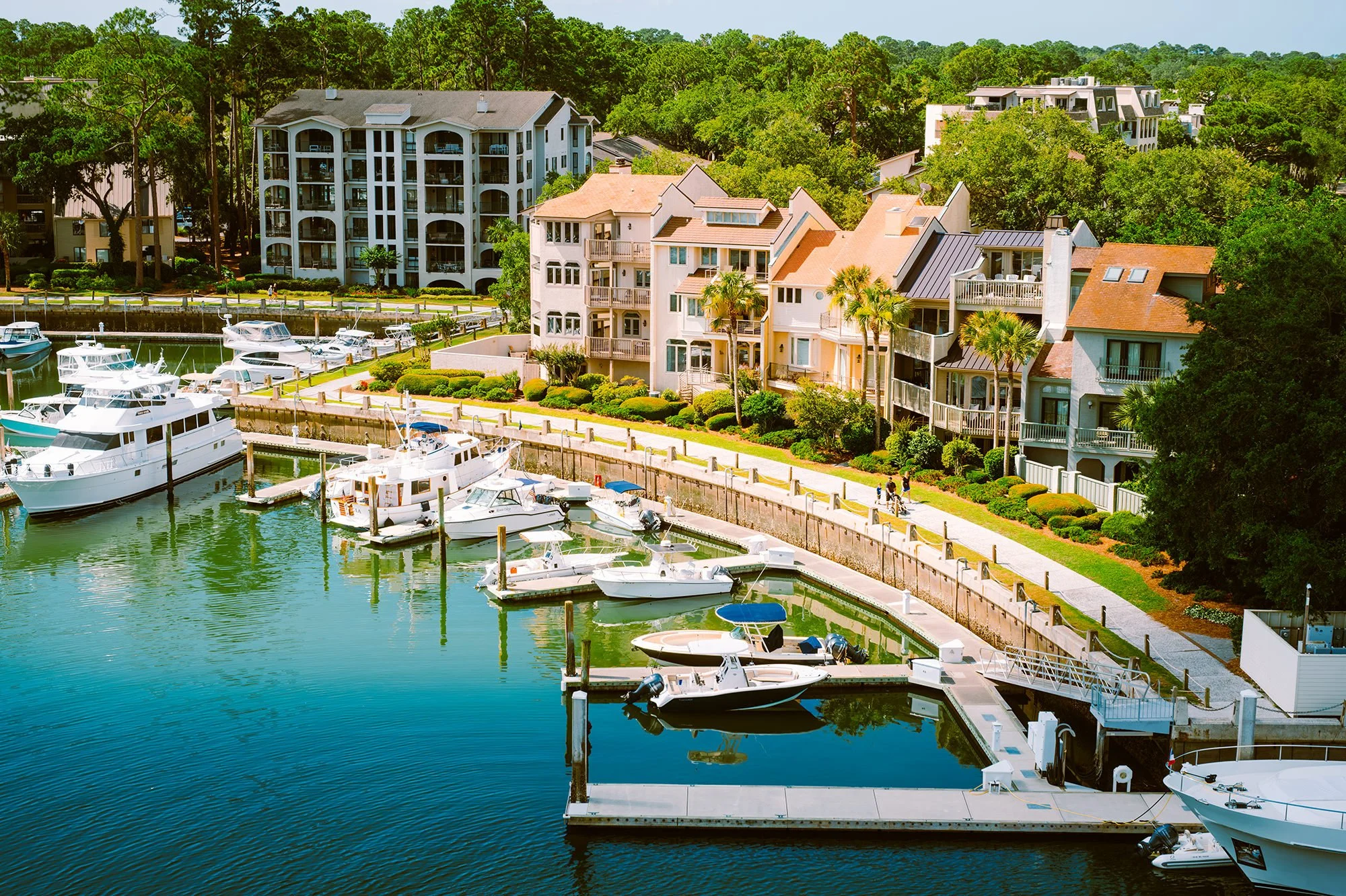 A marina with several boats docked along a waterfront, overlooking multi-story residential buildings with balconies, surrounded by green trees, and a walkway with people strolling.