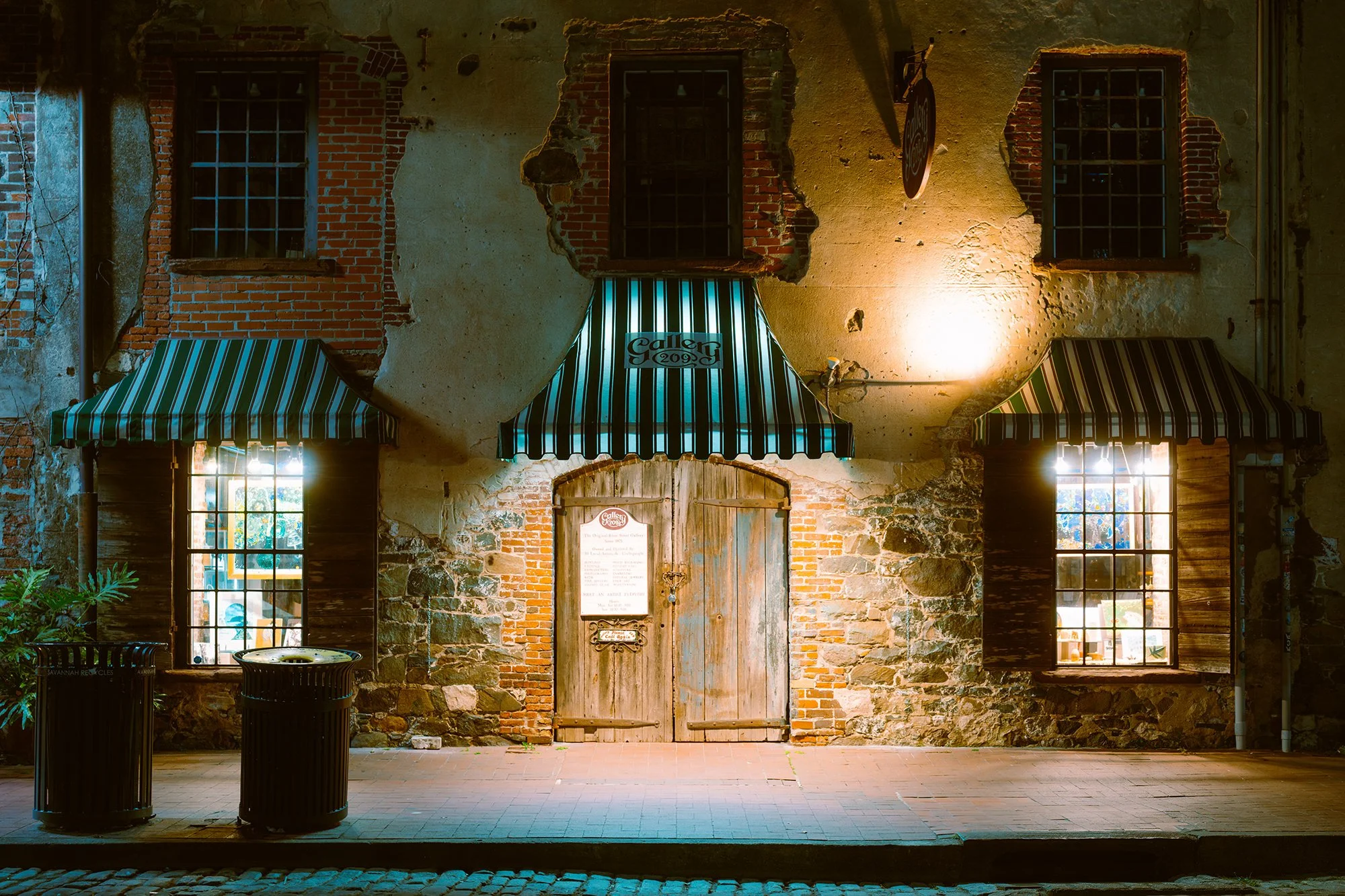Old brick building with three awnings over windows and door, two trash cans in front, warm lighting, and vintage-style wooden double doors.