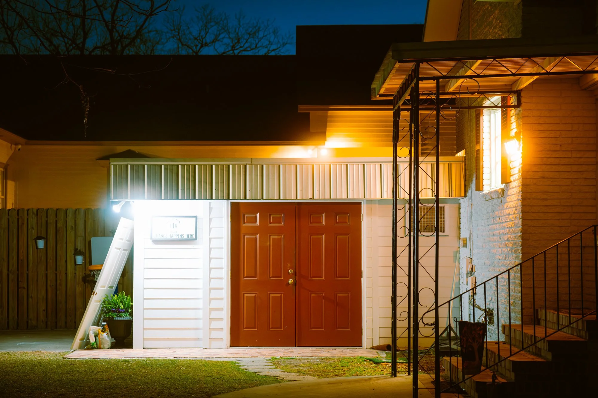 Front view of a house at night with warm exterior lighting, a brown door, white siding, a metal awning, a staircase, plants, and a white sign.