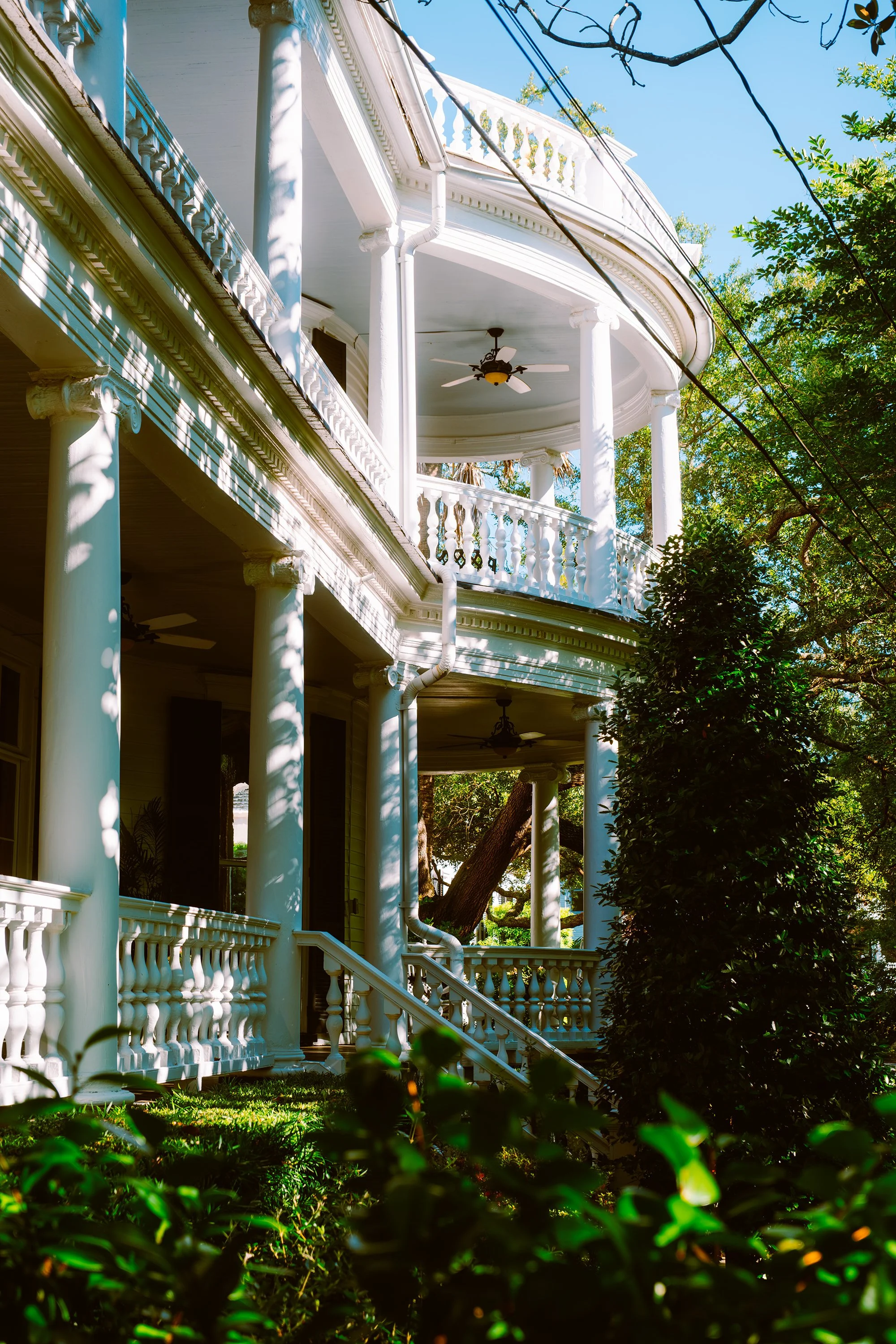 A large, white, multi-story house with columns, a wraparound porch, and a decorative railing. The house has a curved balcony with a ceiling fan and is surrounded by trees and greenery. Sunlight casts shadows on the house.