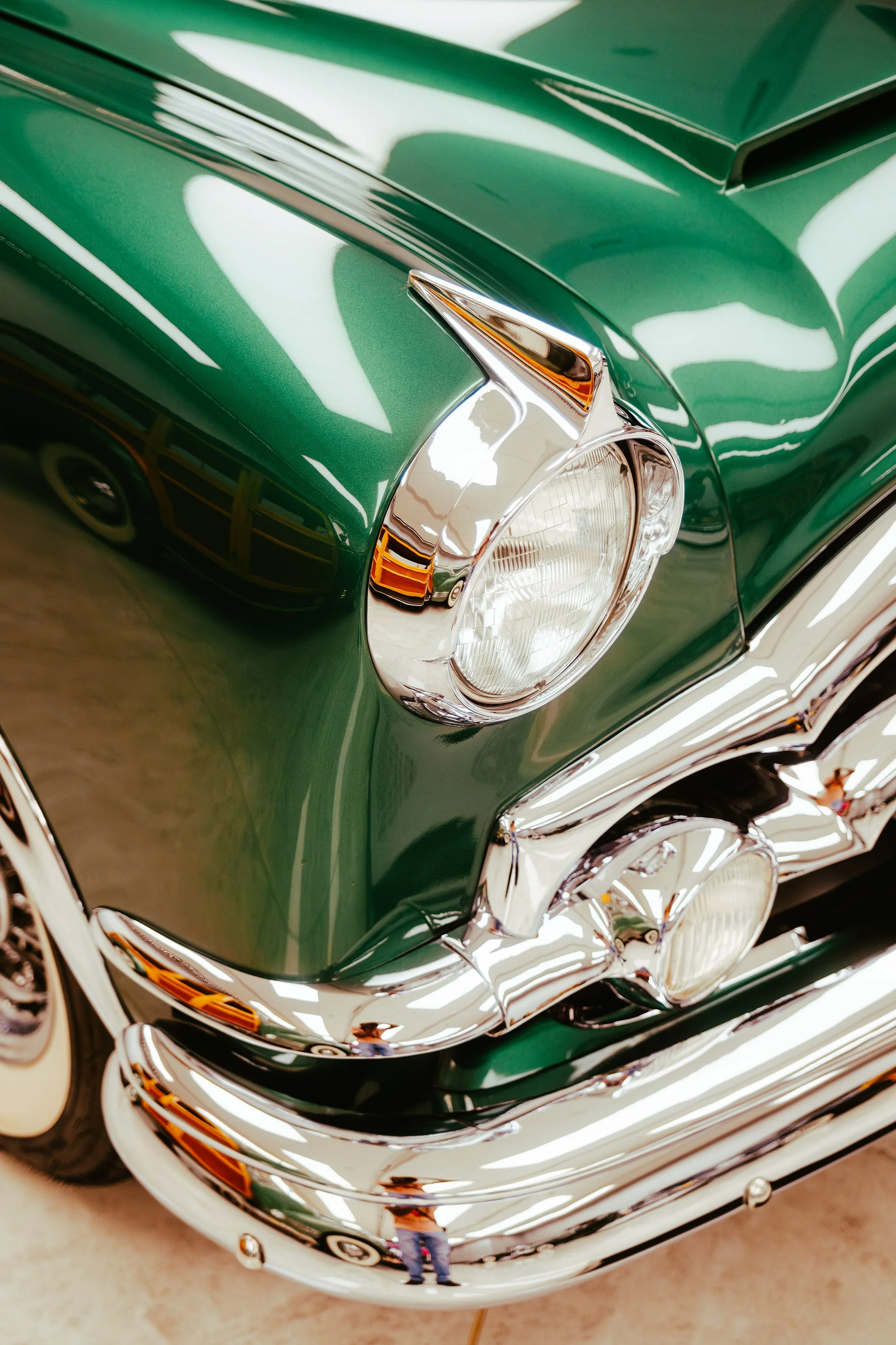 Close-up of a vintage green car's front, showing headlight, chrome bumper, and reflections on the shiny surface.