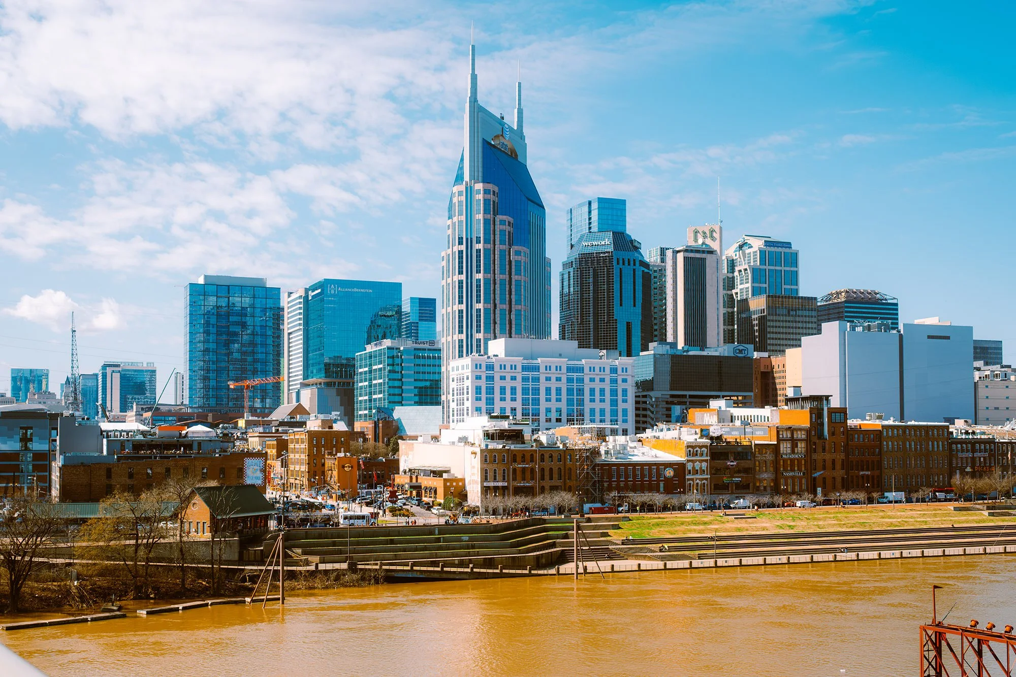City skyline with tall skyscrapers and modern buildings, viewed across a river with brown water.