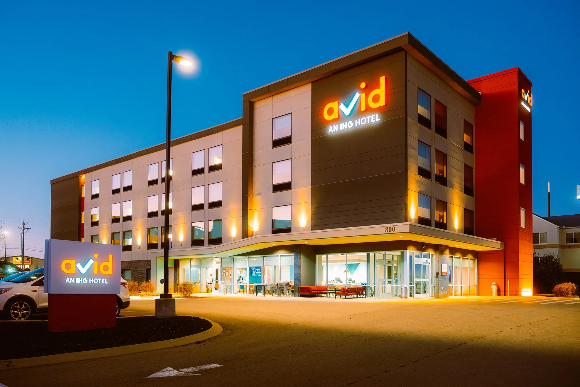Exterior view of a modern name brand hotel at dusk, featuring a brightly lit sign reading 'avid AN IHG HOTEL', with several windows and parking lot in front.