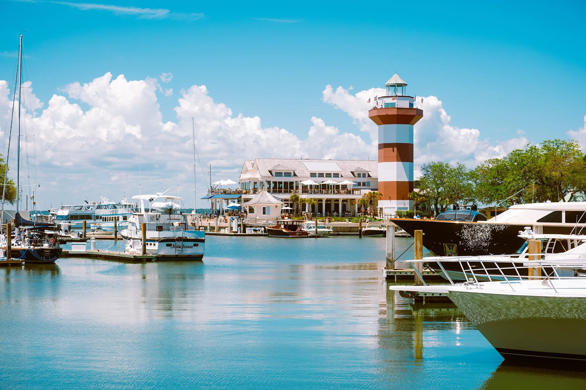 Marina with docked boats, a lighthouse with red, white, and brown stripes, and a waterfront restaurant under a partly cloudy sky.