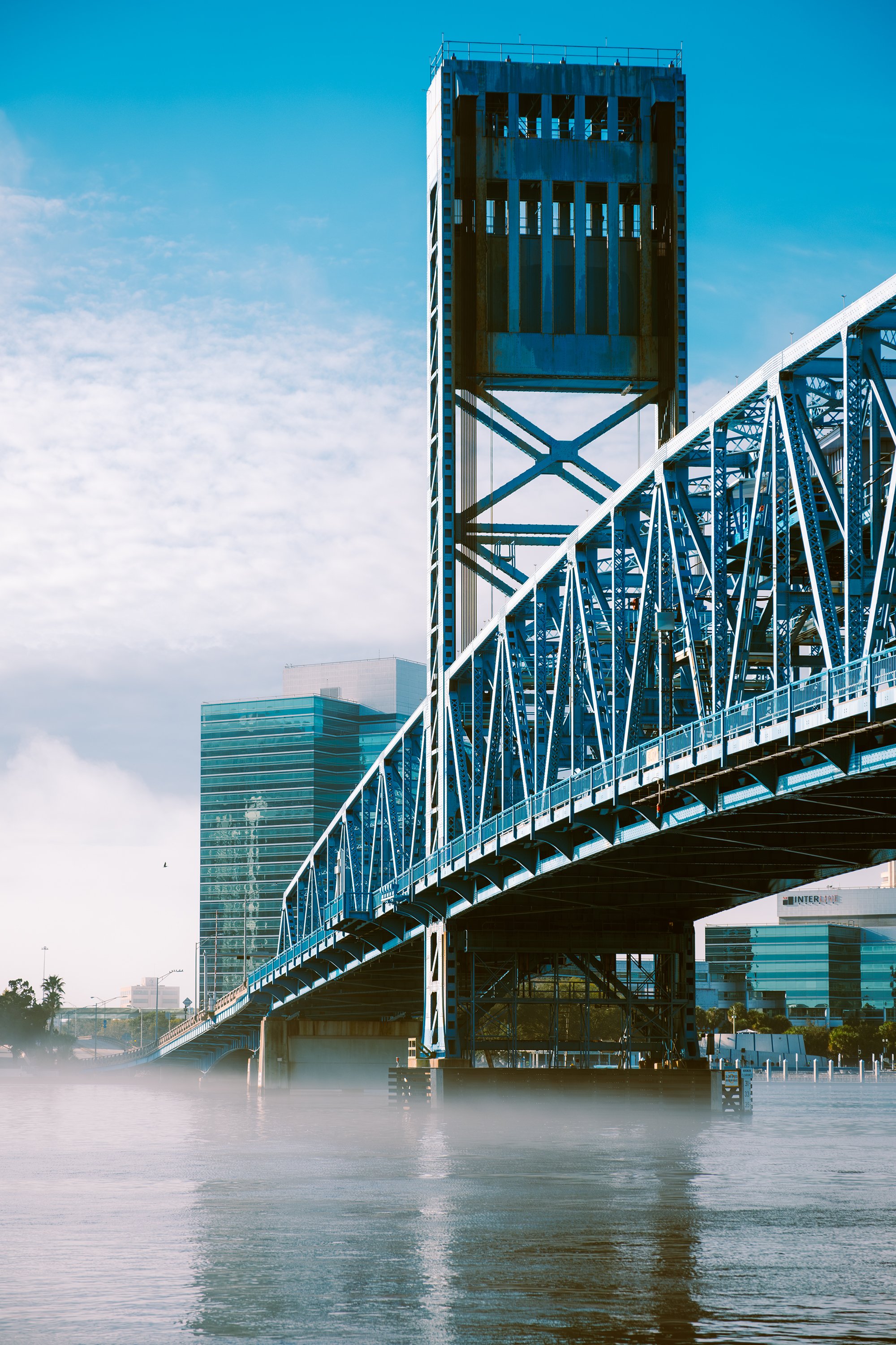 A blue steel bridge over a body of water with city buildings in the background and mist on the water's surface.