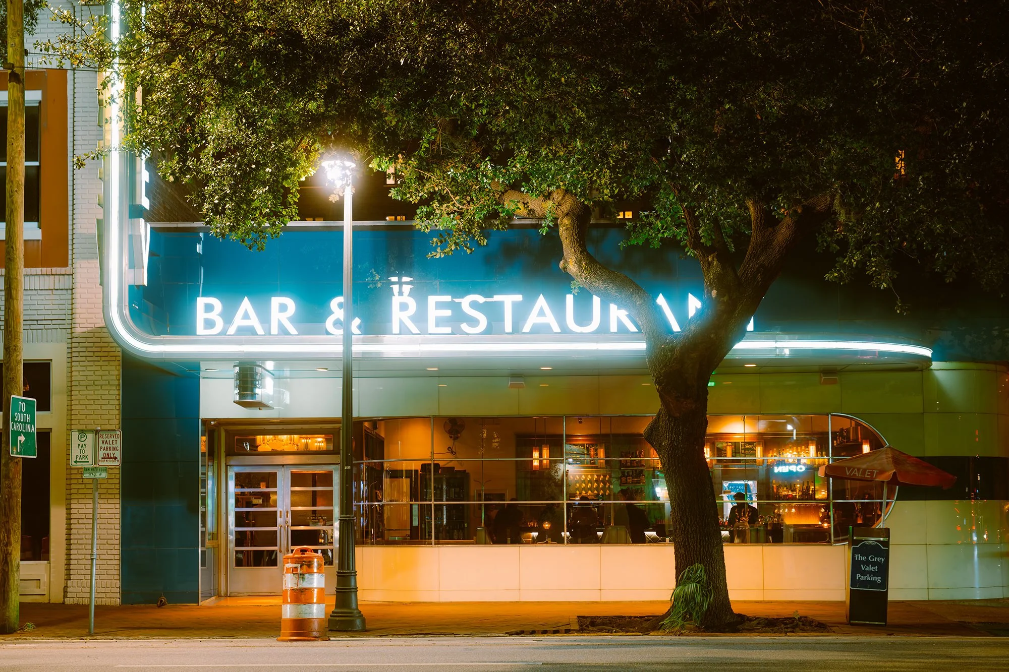 Nighttime view of a bar and restaurant with a neon sign and large front window, with a tree and streetlamp in front.