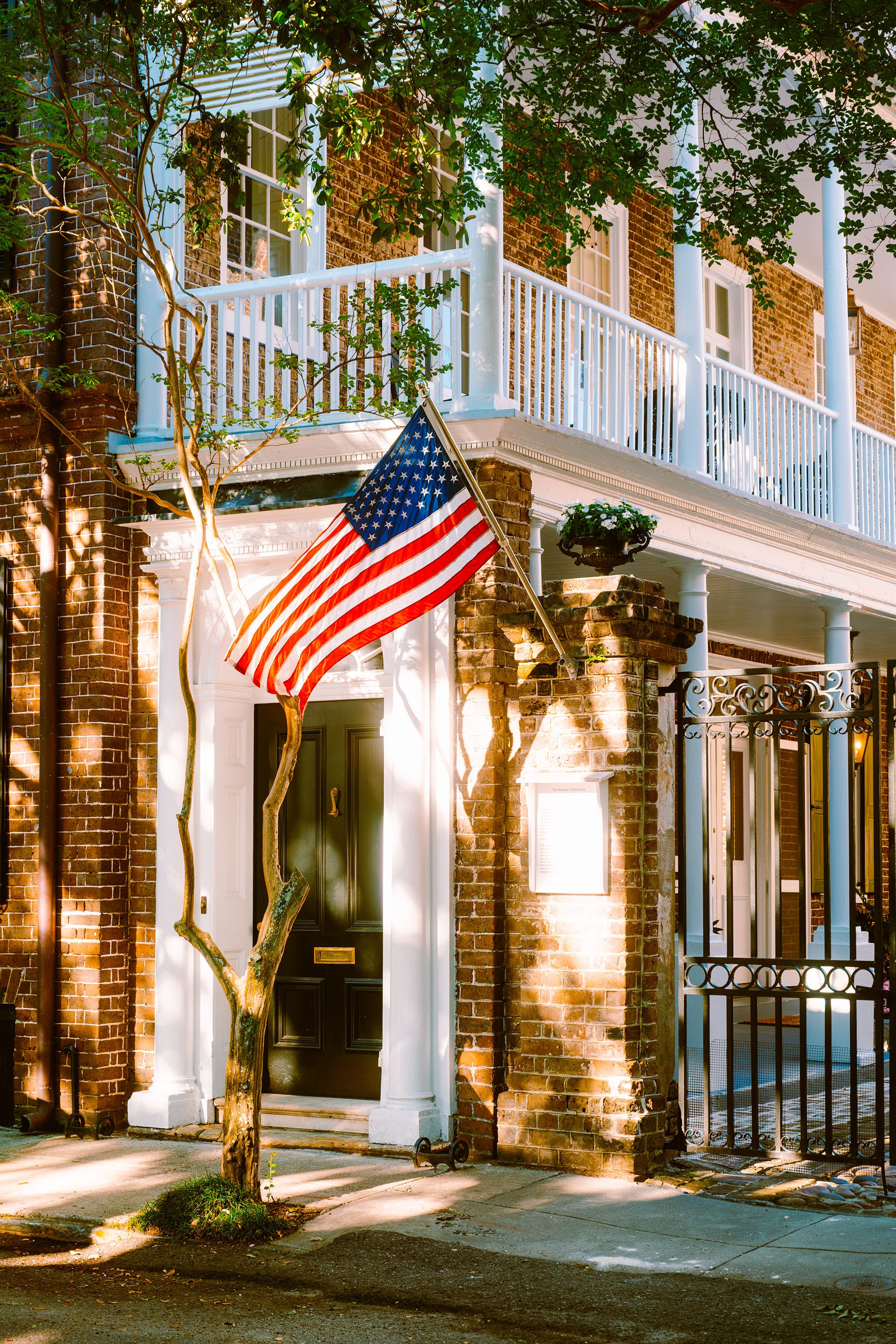 A brick building with white trim and a black front door. An American flag is mounted on the front wall near the door. There is a small tree with twisted branches in front of the door and a balcony with a white railing above.