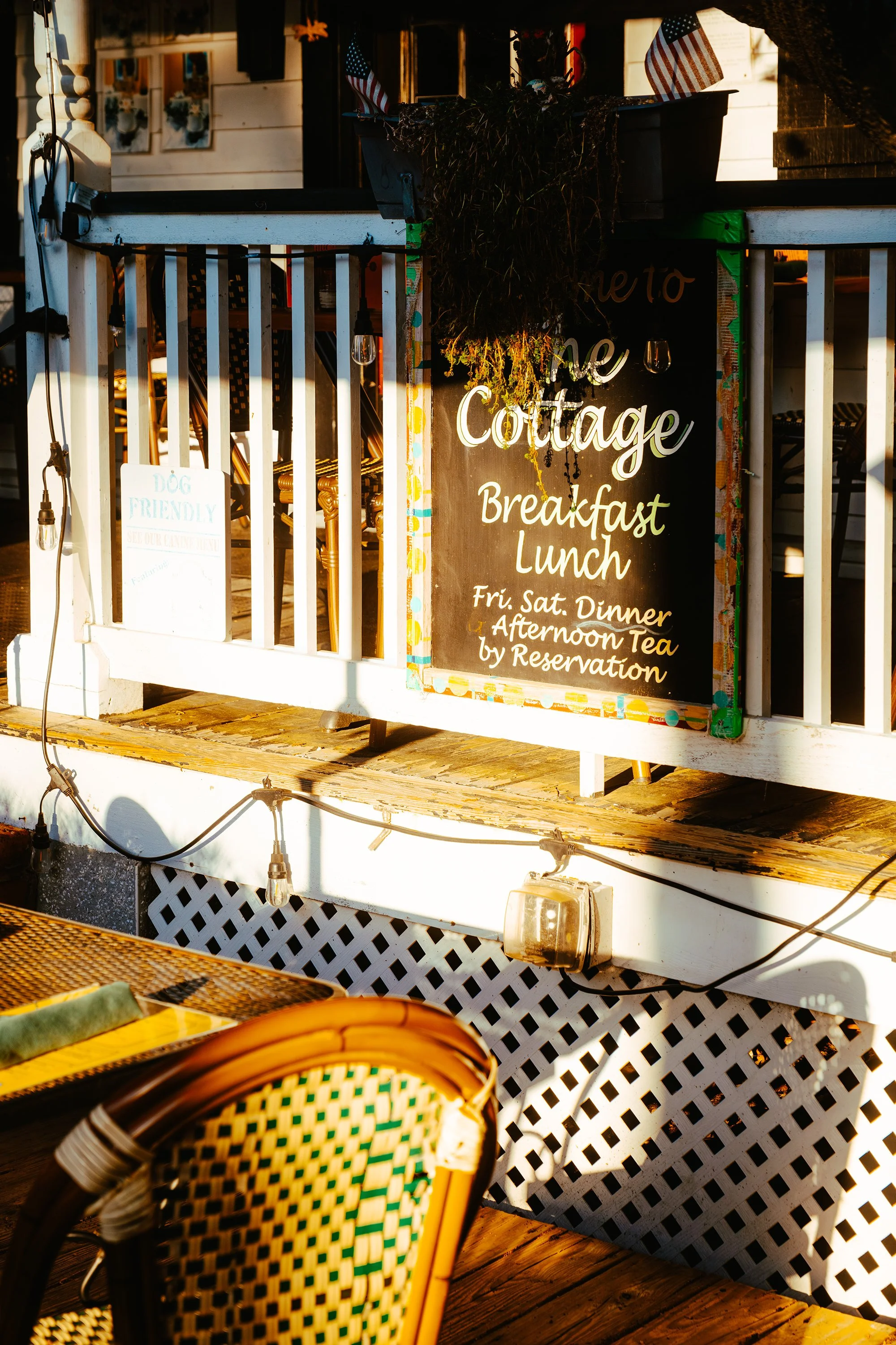 An outdoor restaurant with a blackboard sign listing breakfast, lunch, and dinner hours. There are string lights and American flags, and part of a wooden table and woven chair are visible in the foreground.