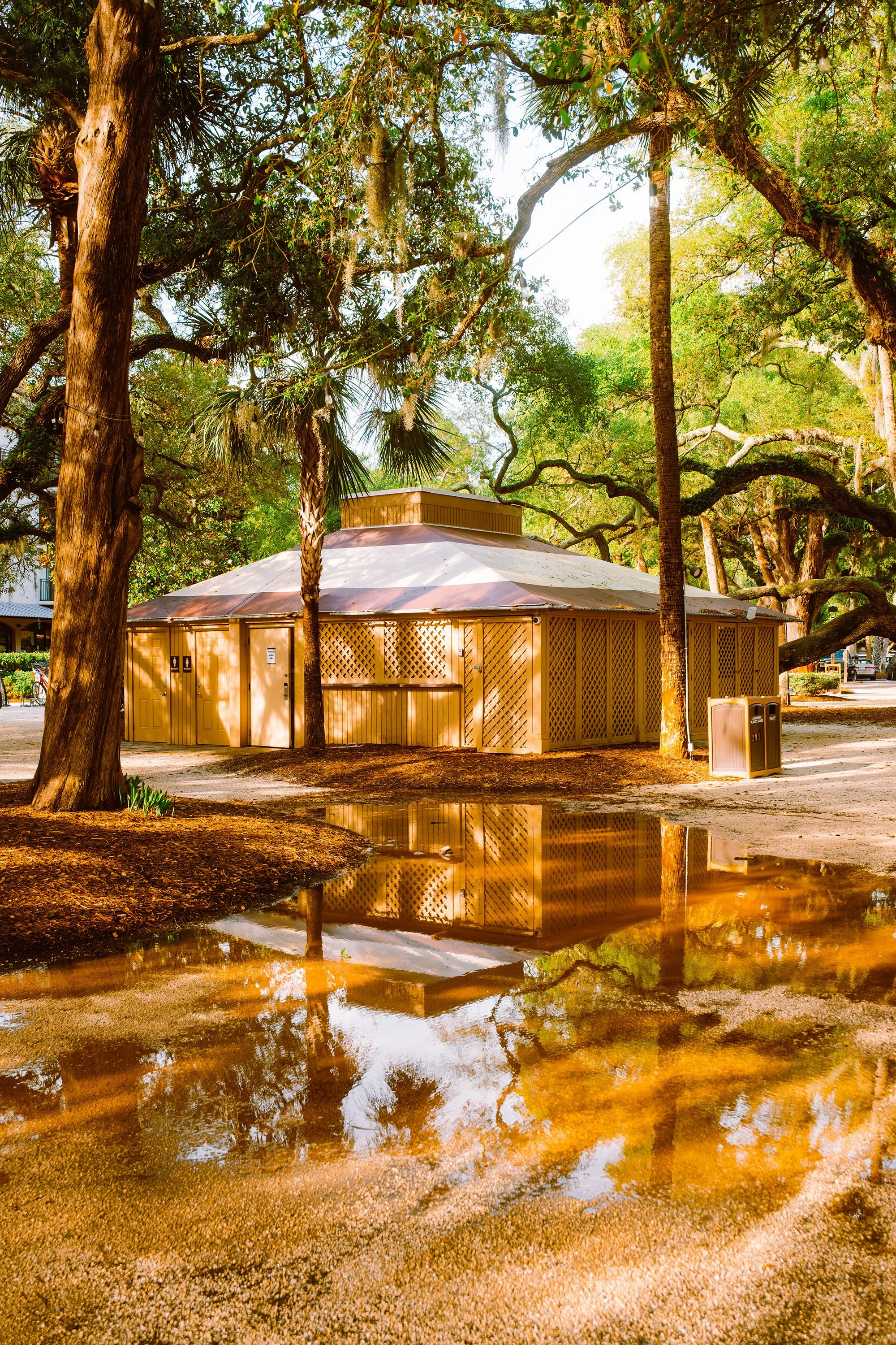 A round wooden pavilion surrounded by large trees with green foliage, reflected in a puddle on the ground.