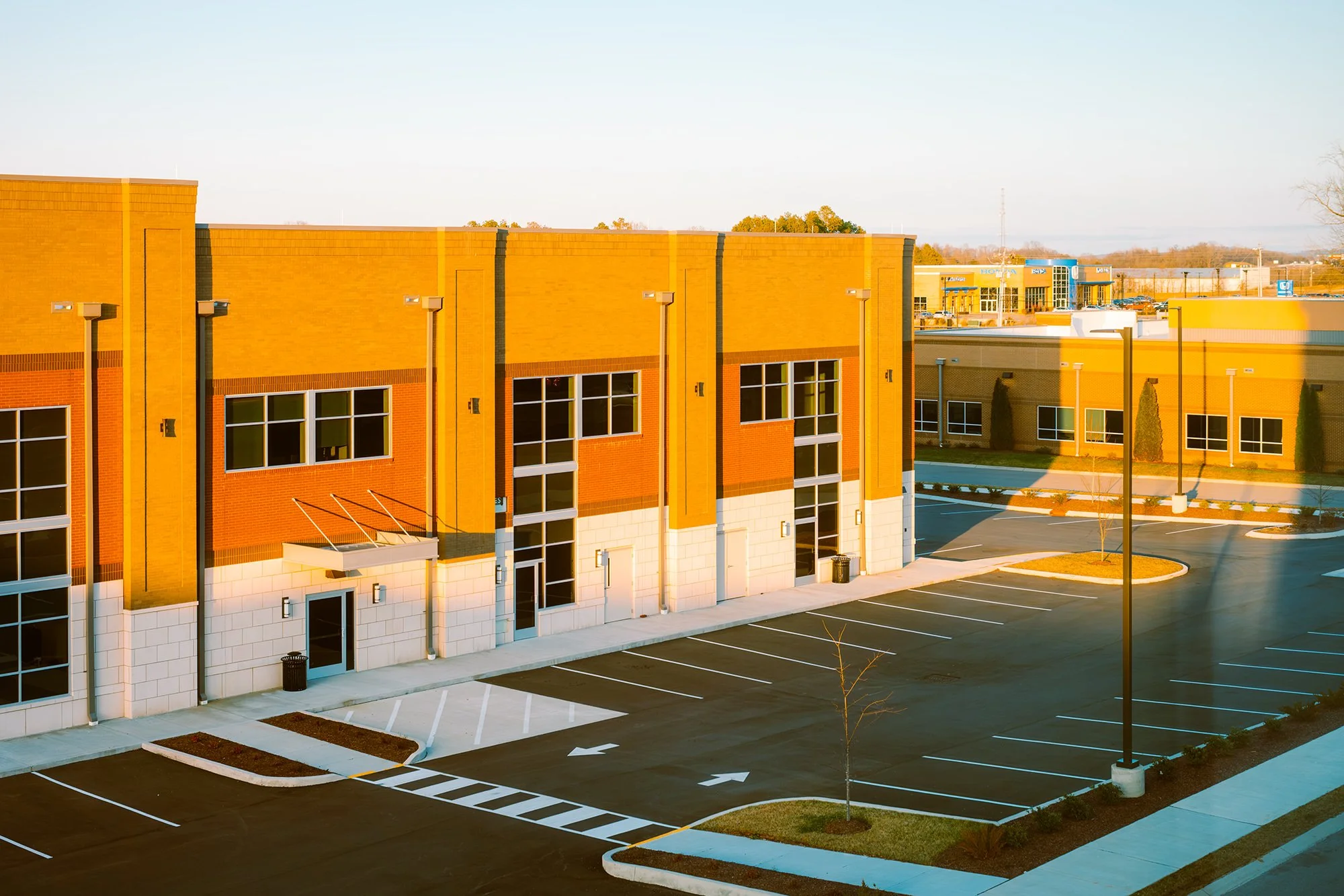 Empty parking lot outside a modern yellow brick building with trees and additional commercial buildings in the background.