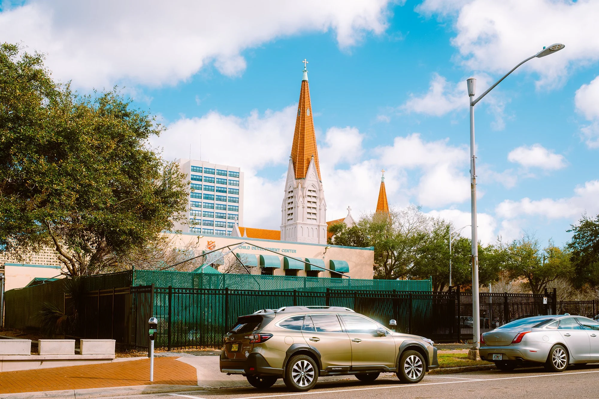 Street scene with parked cars, trees, streetlamp, and a church with tall steeples in the background under a partly cloudy sky.