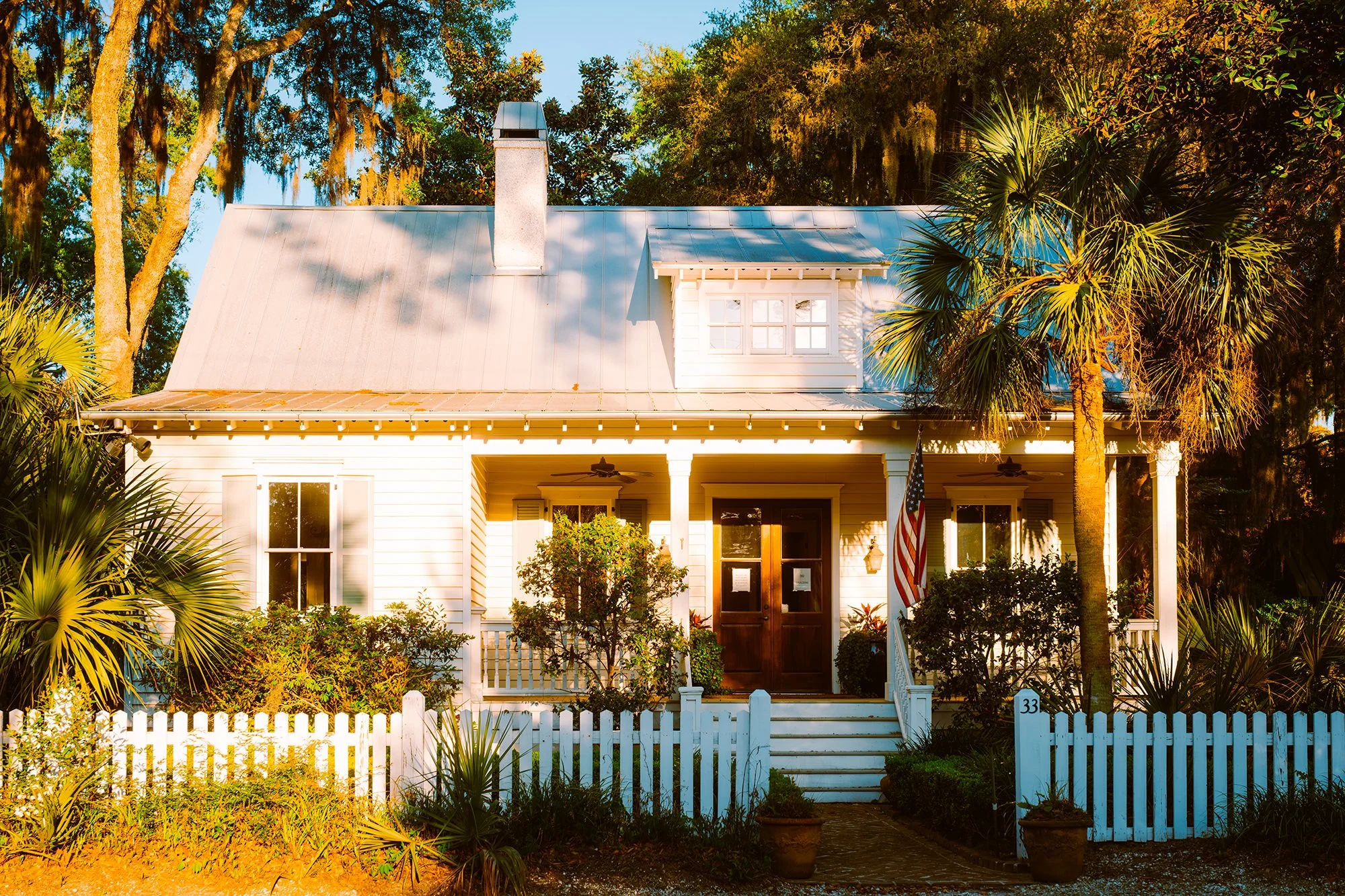 A white house with a metal roof, surrounded by trees and bushes, featuring a front porch with steps, an American flag, and a white picket fence.