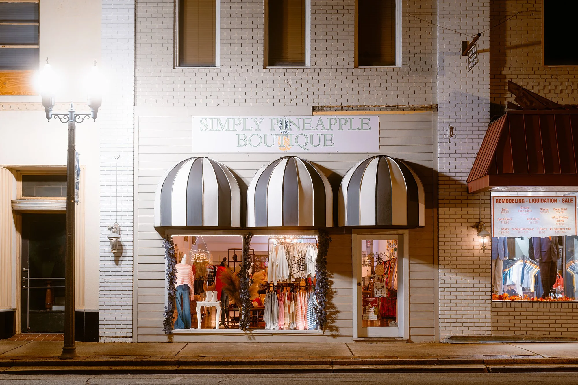 Night view of a boutique clothing store named 'Simply Pineapple Boutique' with striped black and white awnings, mannequins dressed in clothing inside the window, and a lit 'OPEN' sign. To the right, a sign advertises remodeling, liquidation, and sale