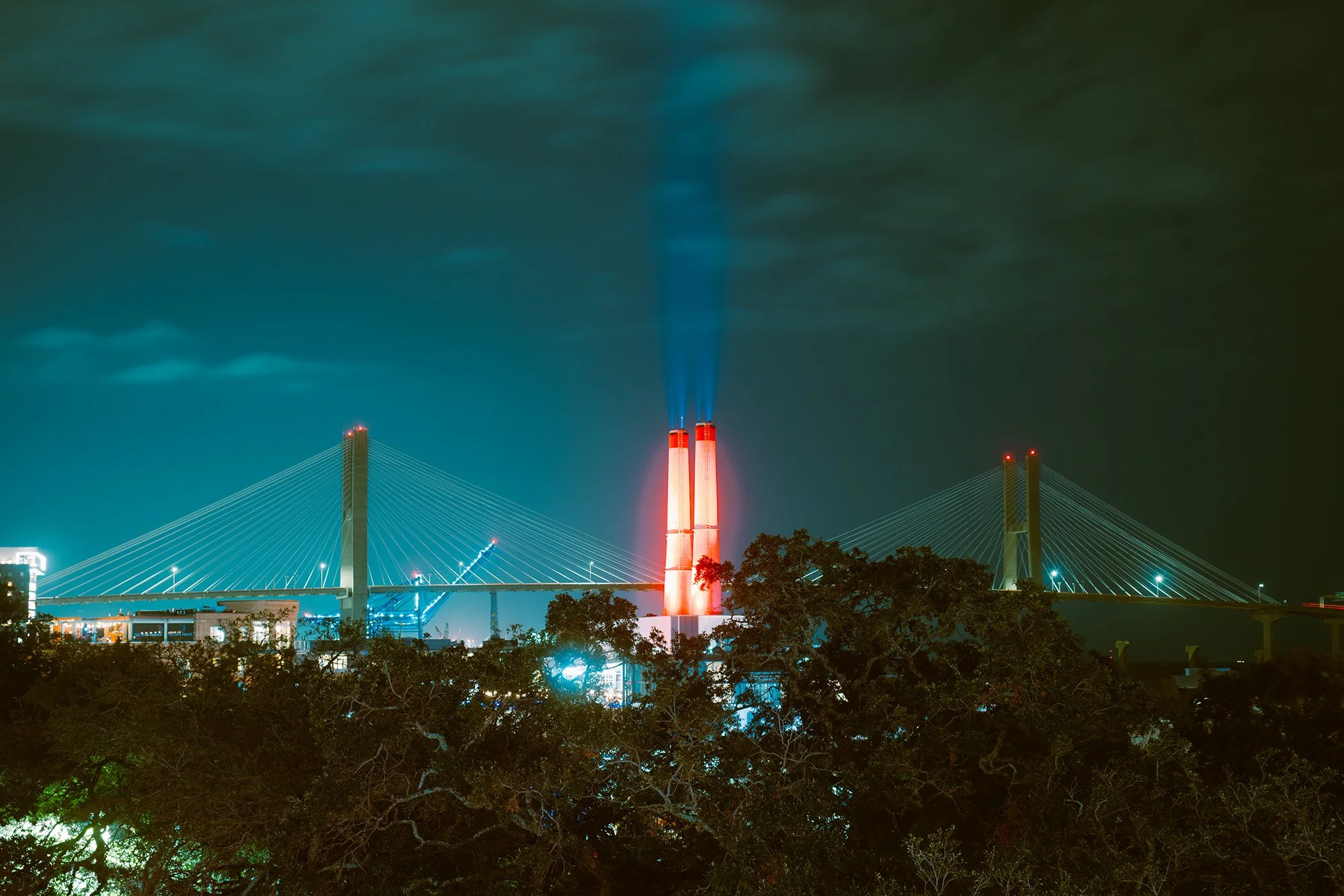 Night scene of a city with a bridge and two tall smokestacks emitting blue smoke, illuminated with red and blue lights, and surrounded by dark trees.