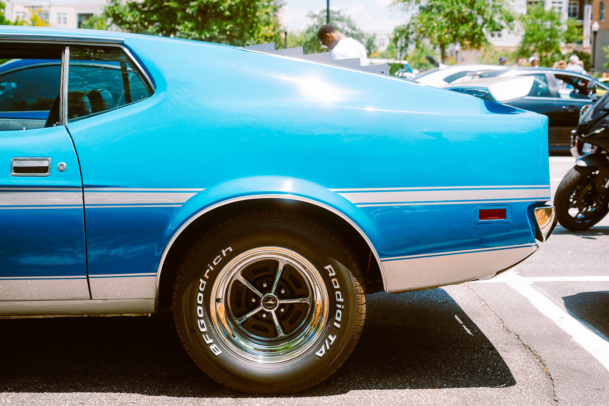Close-up of a blue vintage car parked in a parking lot with other vehicles and people in the background.