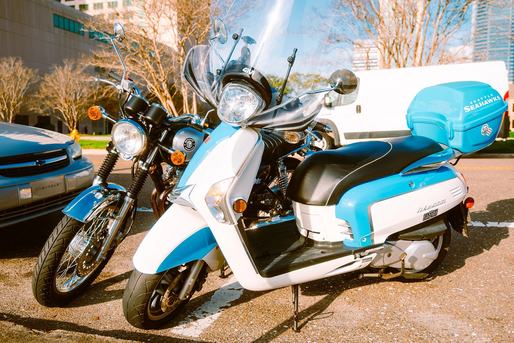Blue and white scooter parked in a lot with a black motorcycle nearby and cars and trees in the background.