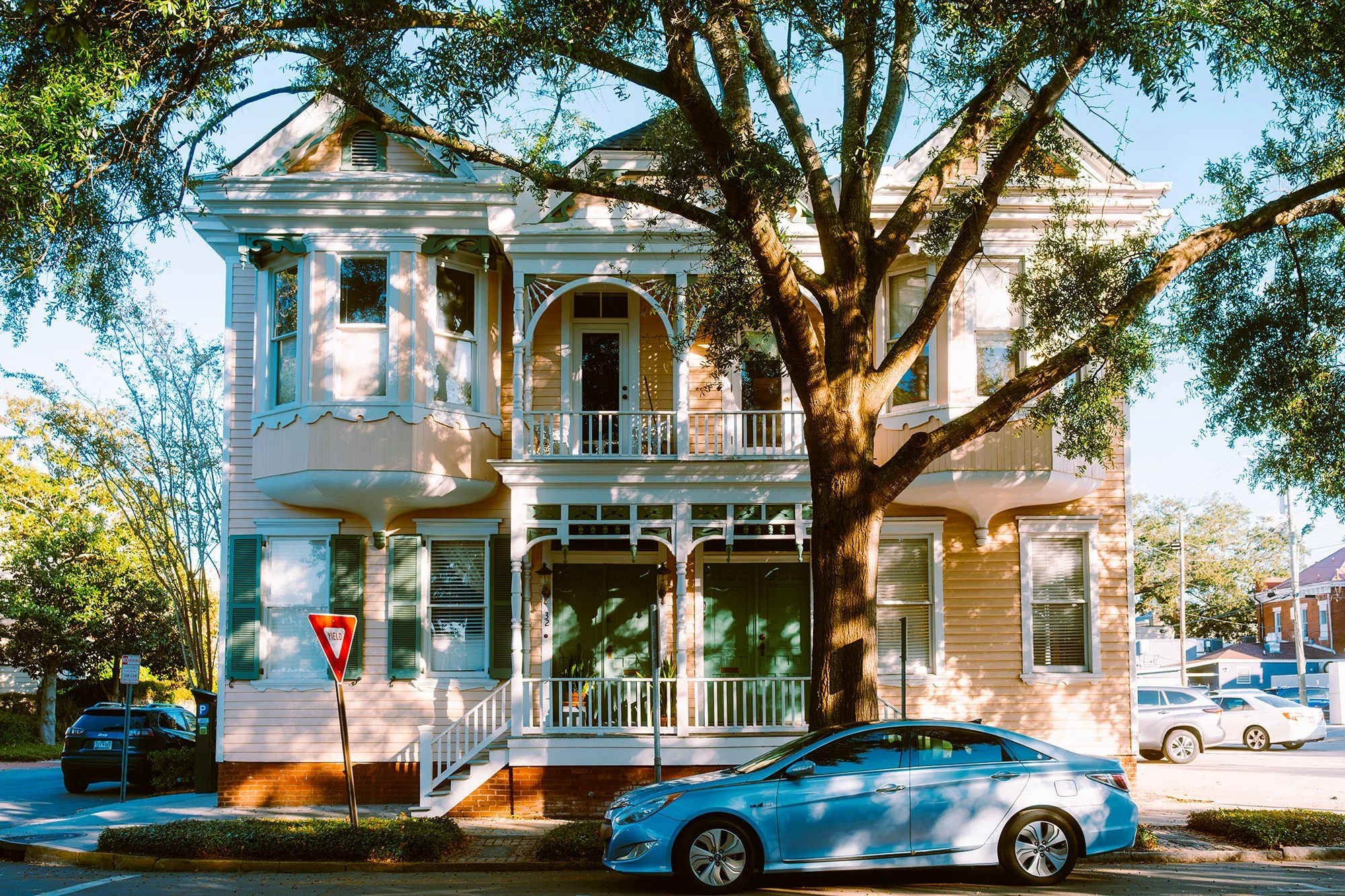 A two-story Victorian-style house painted in pale yellow with white trim, large windows, a small front porch, and a prominent tree in the front yard, with a silver car parked in front.