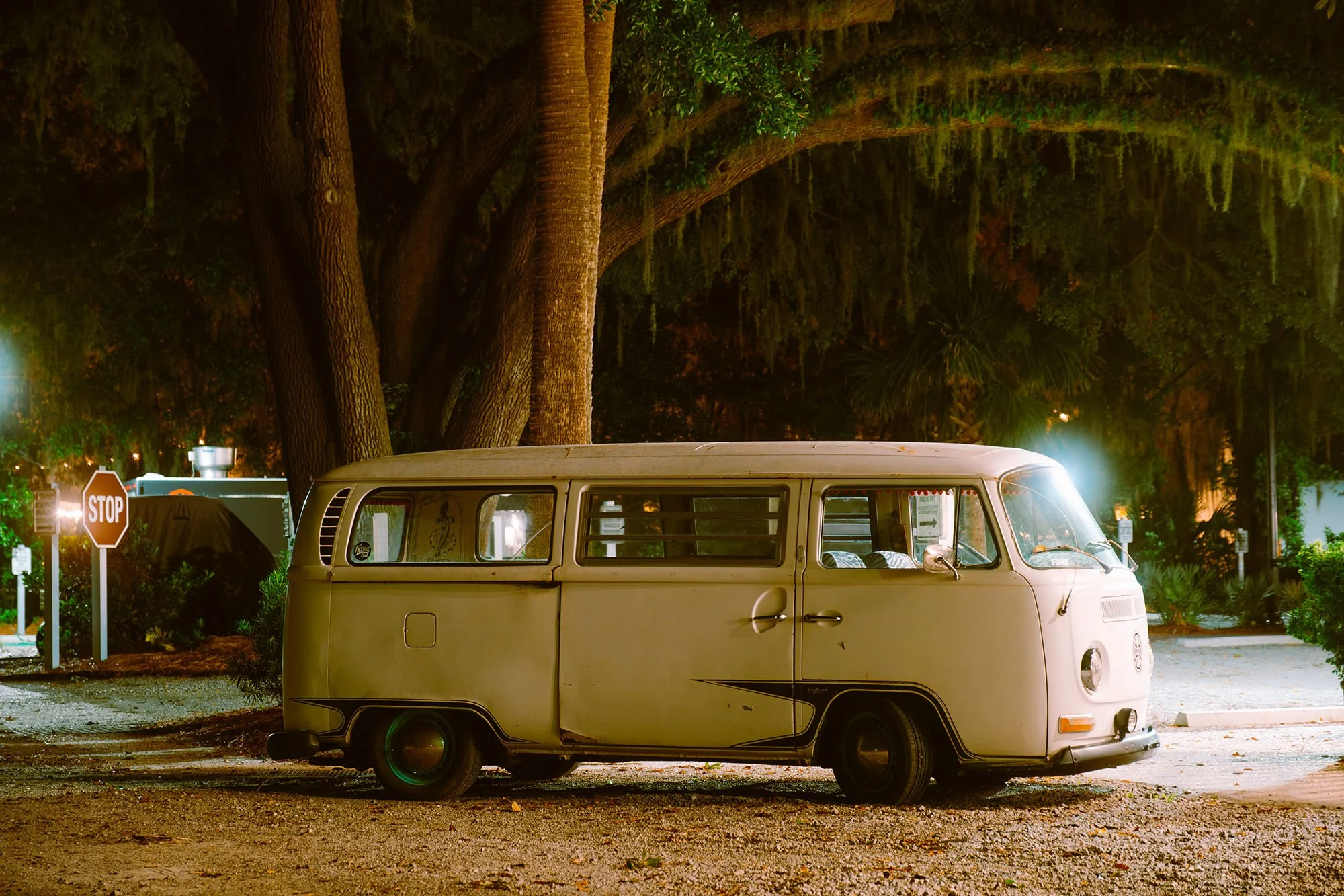 A vintage Volkswagen camper van parked on a street at night, illuminated by streetlights, with large trees in the background and a stop sign nearby.