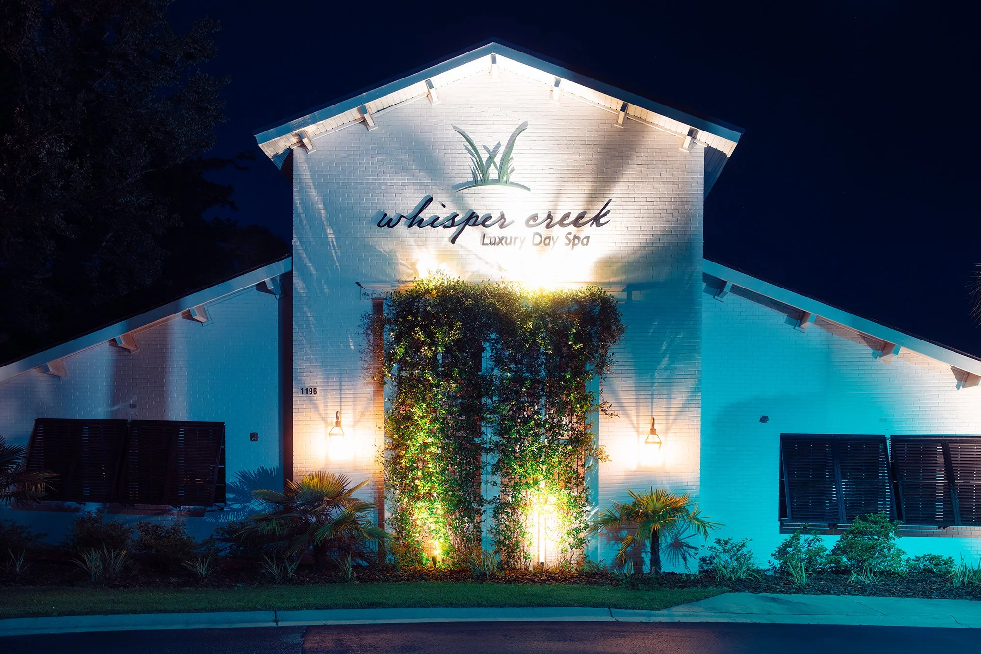 Front of a white building labeled 'whisper creek Luxury Day Spa' illuminated at night, with greenery and decorative lighting.