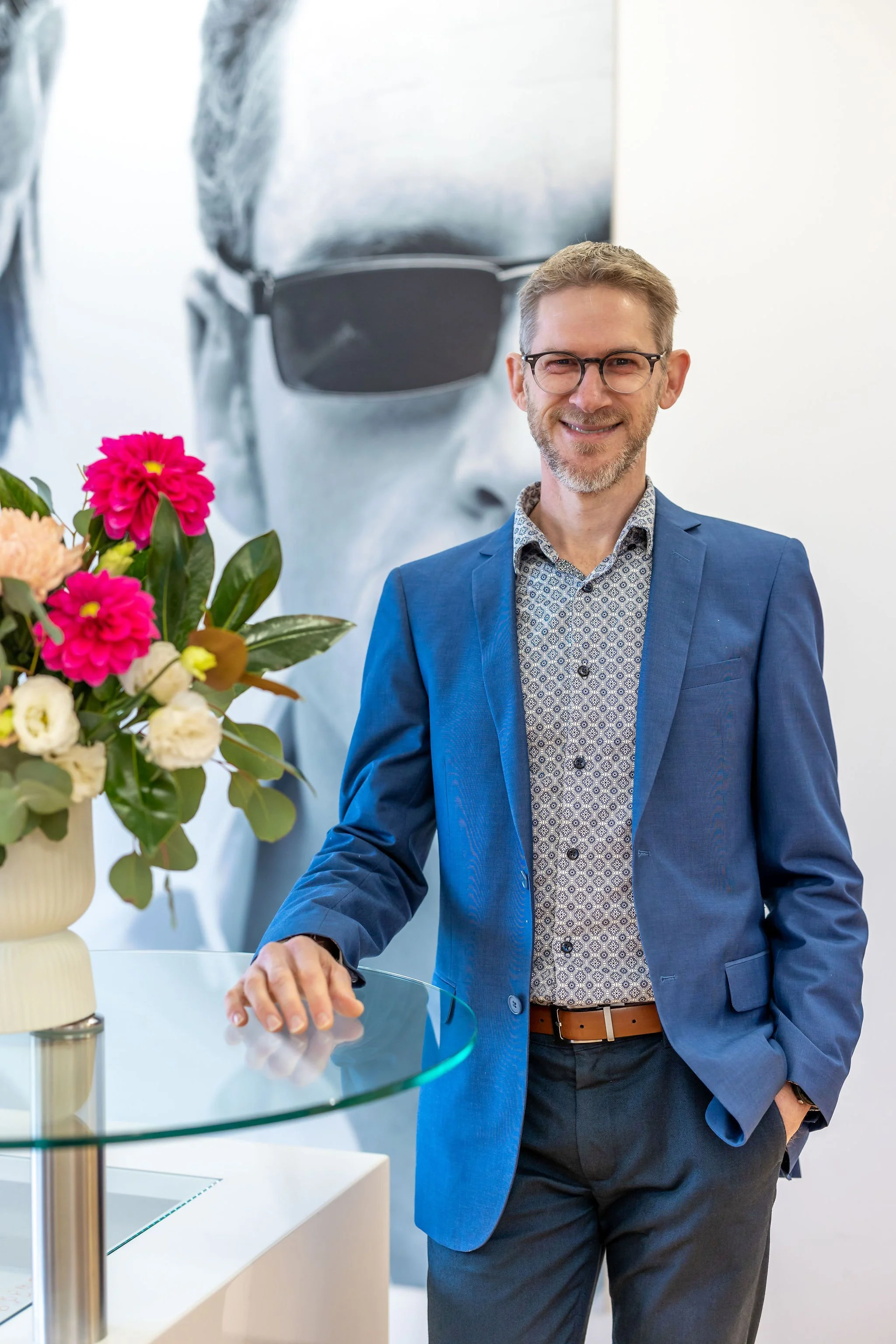 Man in blue suit standing next to a glass table with a vase of flowers, large black and white poster of person in sunglasses in the background.