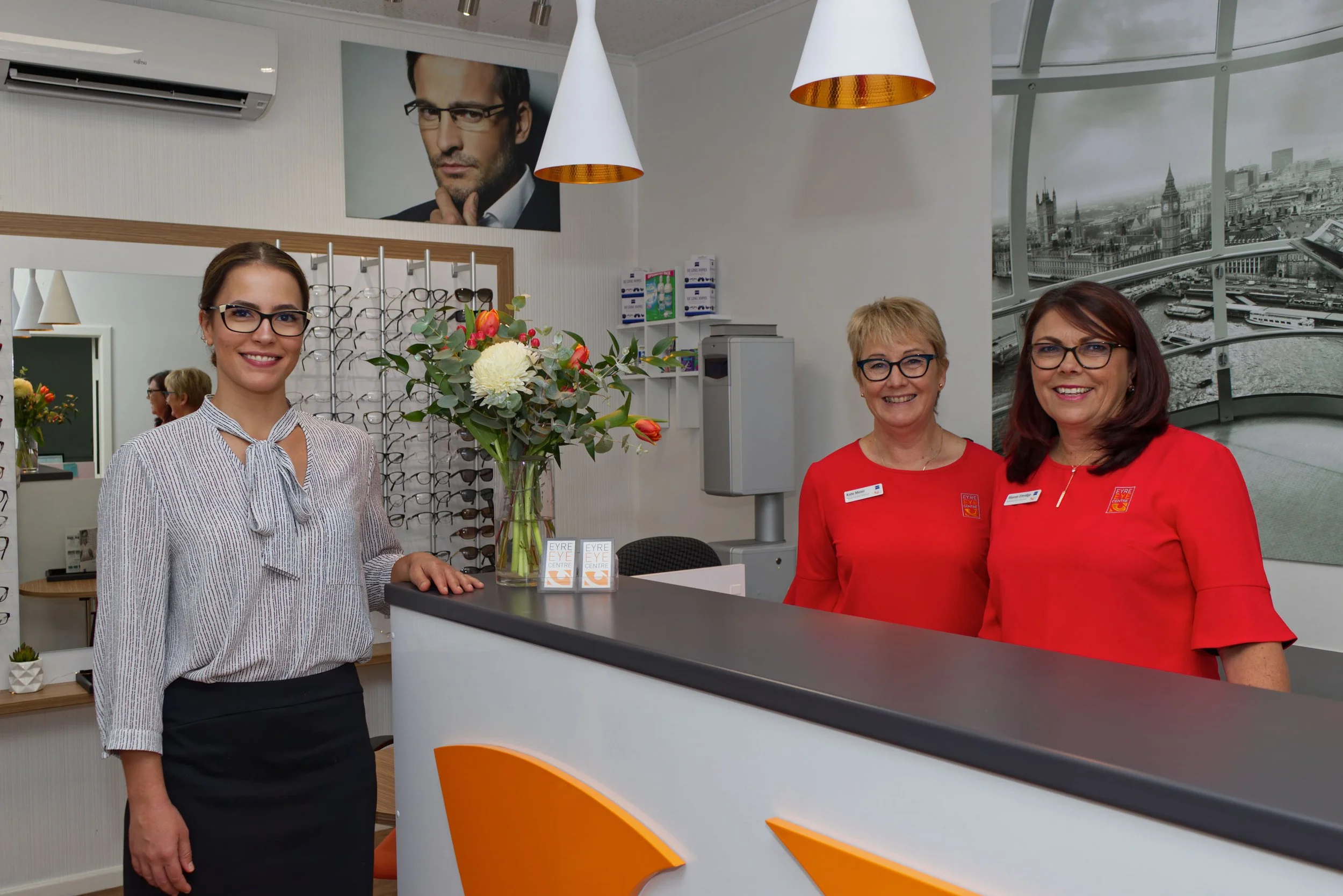 Three women standing at the reception desk of an optical store, two wearing red uniforms. Glasses display in the background and a large poster of a man wearing glasses on the wall. A vase of flowers is on the counter.