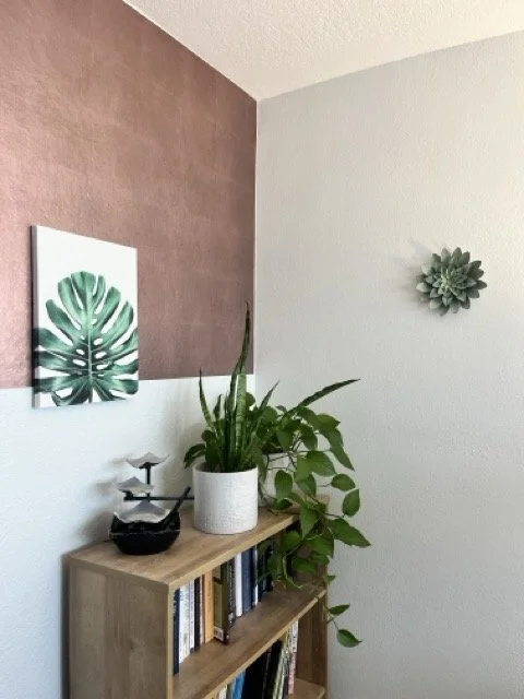 Interior corner of a room with a wooden bookshelf, houseplants, framed leaf art, and a wall decoration.