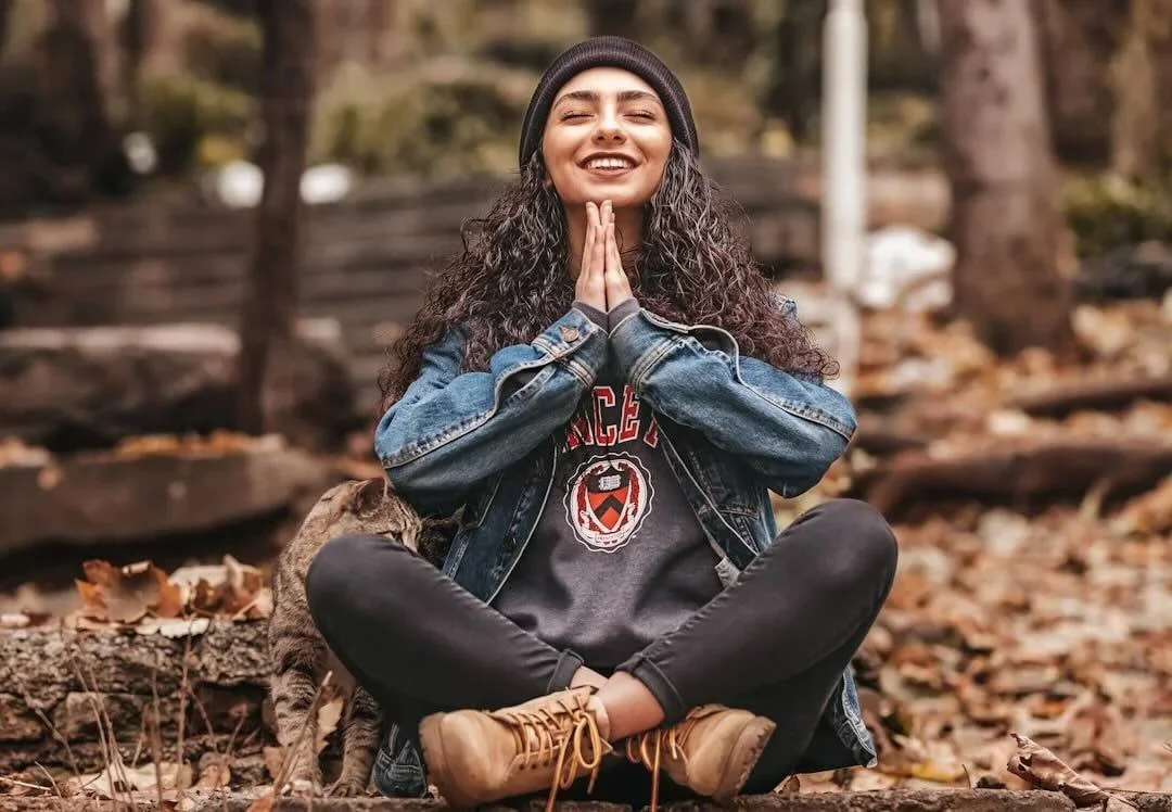 A young woman with curly hair, wearing a black beanie, denim jacket, and casual clothing, is sitting outdoors on a fallen log surrounded by autumn leaves, with her hands pressed together in a prayer position and smiling.