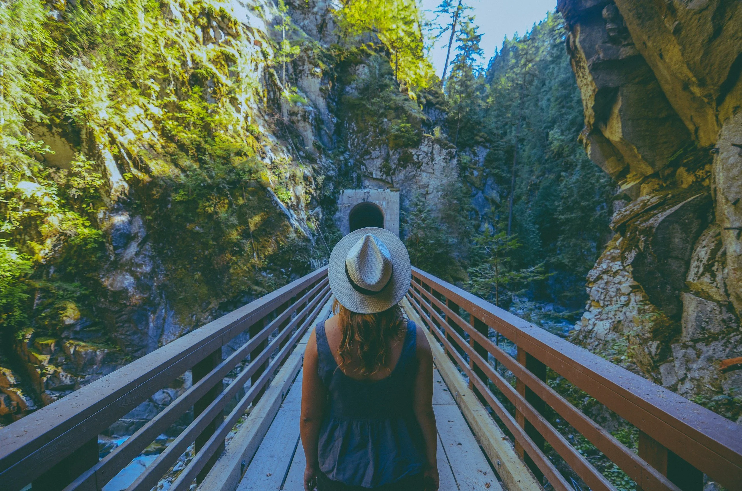 A woman with a hat walking on a wooden bridge in a forested mountain area towards a tunnel entrance.