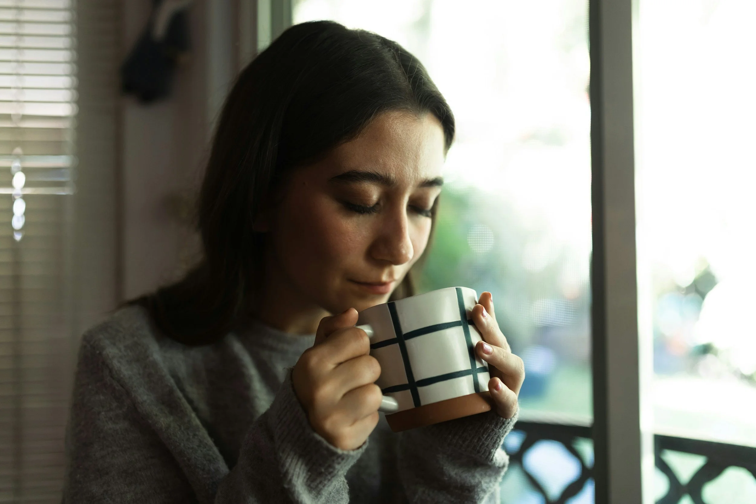A woman with dark hair and a gray sweater holding a white mug with black grid lines near her face, standing by a window.